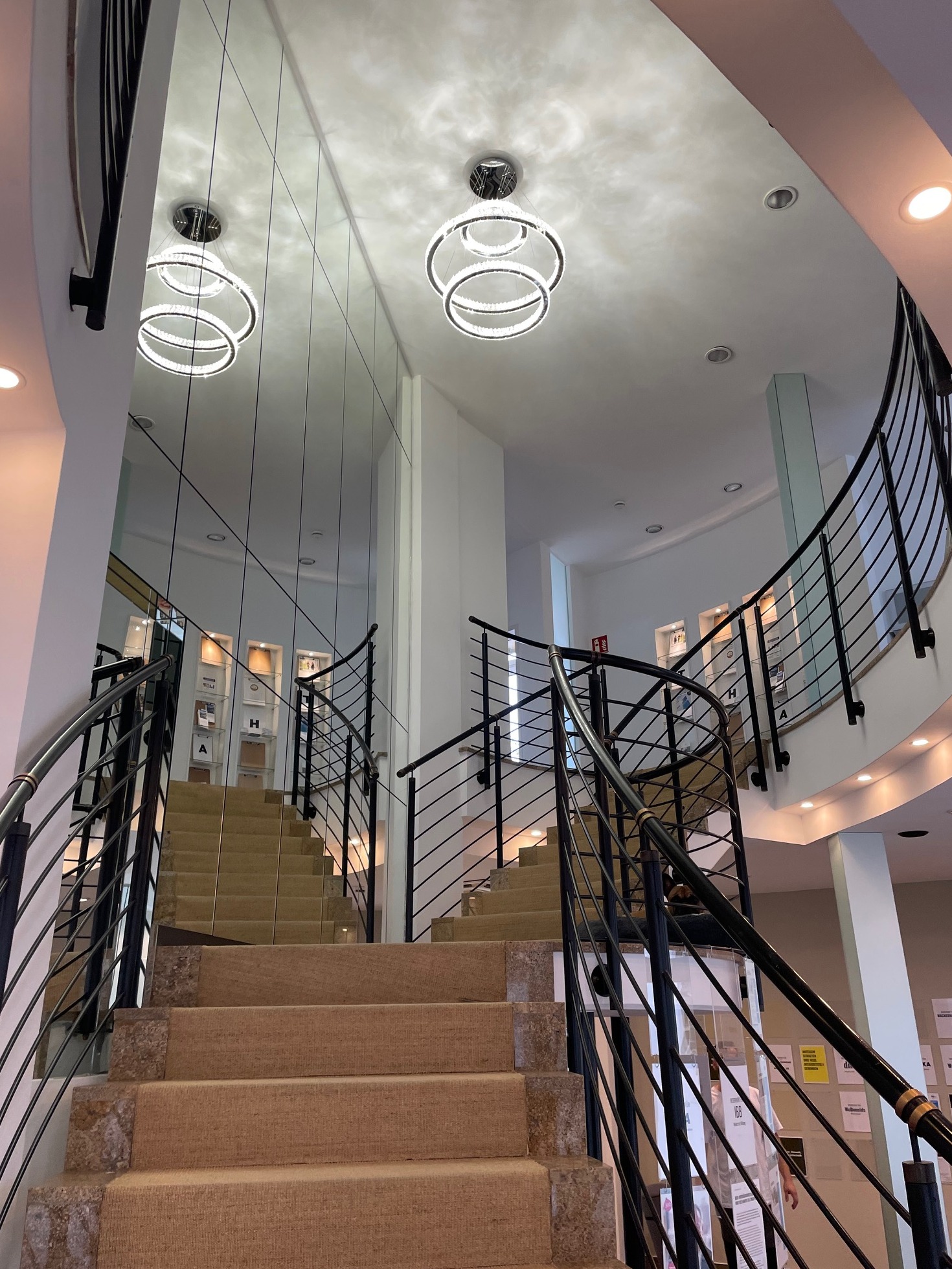Wnętrze budynku użyteczności publicznej ze schodami wyłożonymi wykładziną, czarnymi metalowymi balustradami, lustrem na ścianie oraz nowoczesnymi, okrągłymi żyrandolami LED.