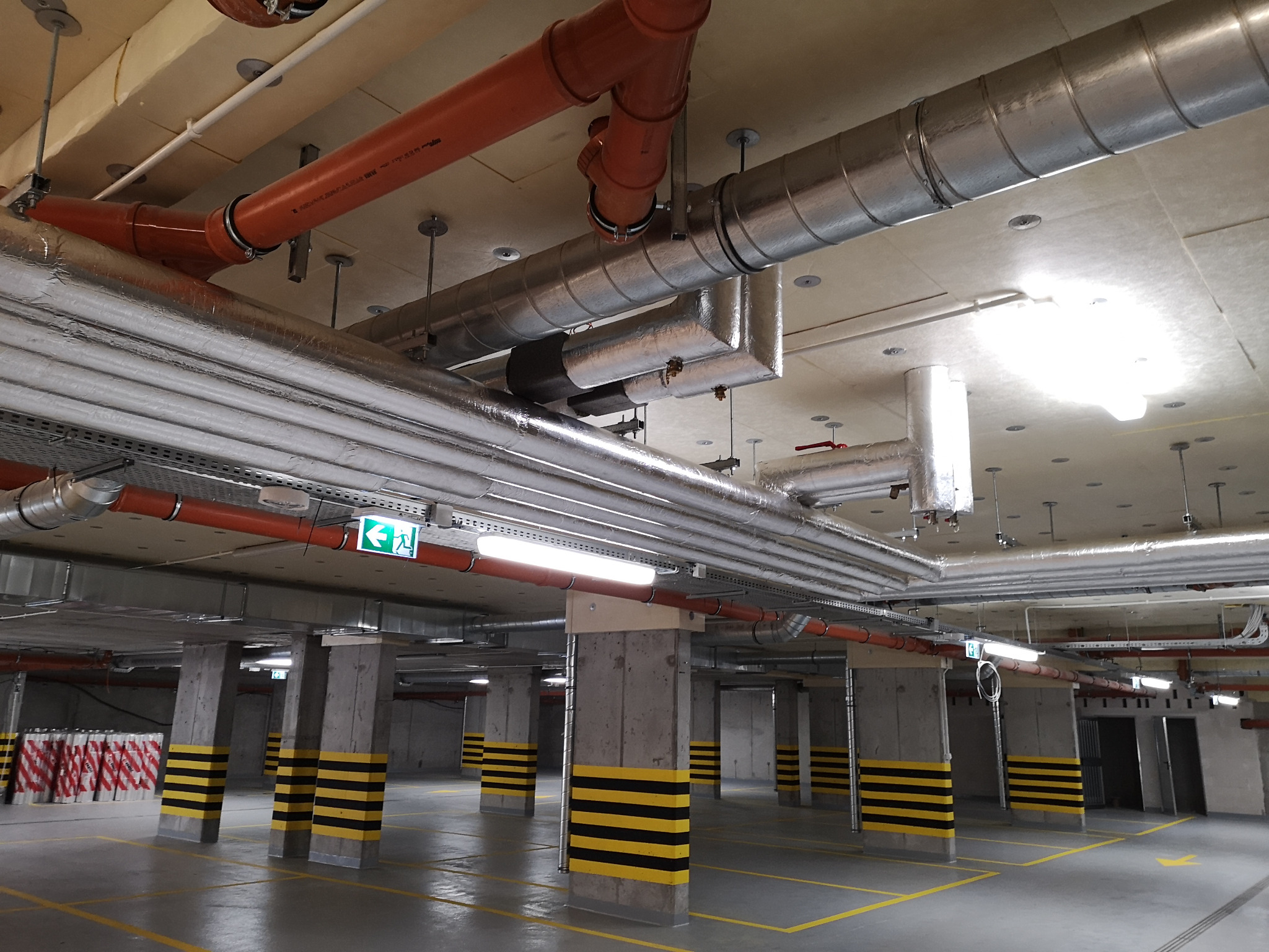 Instalacje wentylacyjne i rury w garażu podziemnym z betonowymi słupami pomalowanymi w czarno-żółte pasy.