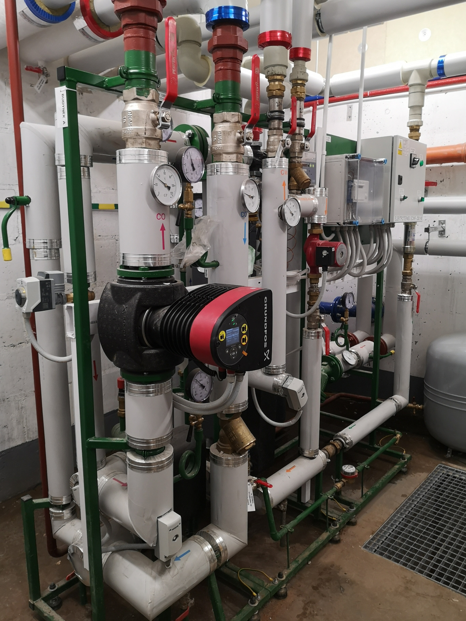 Rozbudowana instalacja grzewcza z pompą obiegową Grundfos, licznymi rurami, zaworami, manometrami i skrzynką elektryczną, oznaczona etykietą 'Budynek A'.