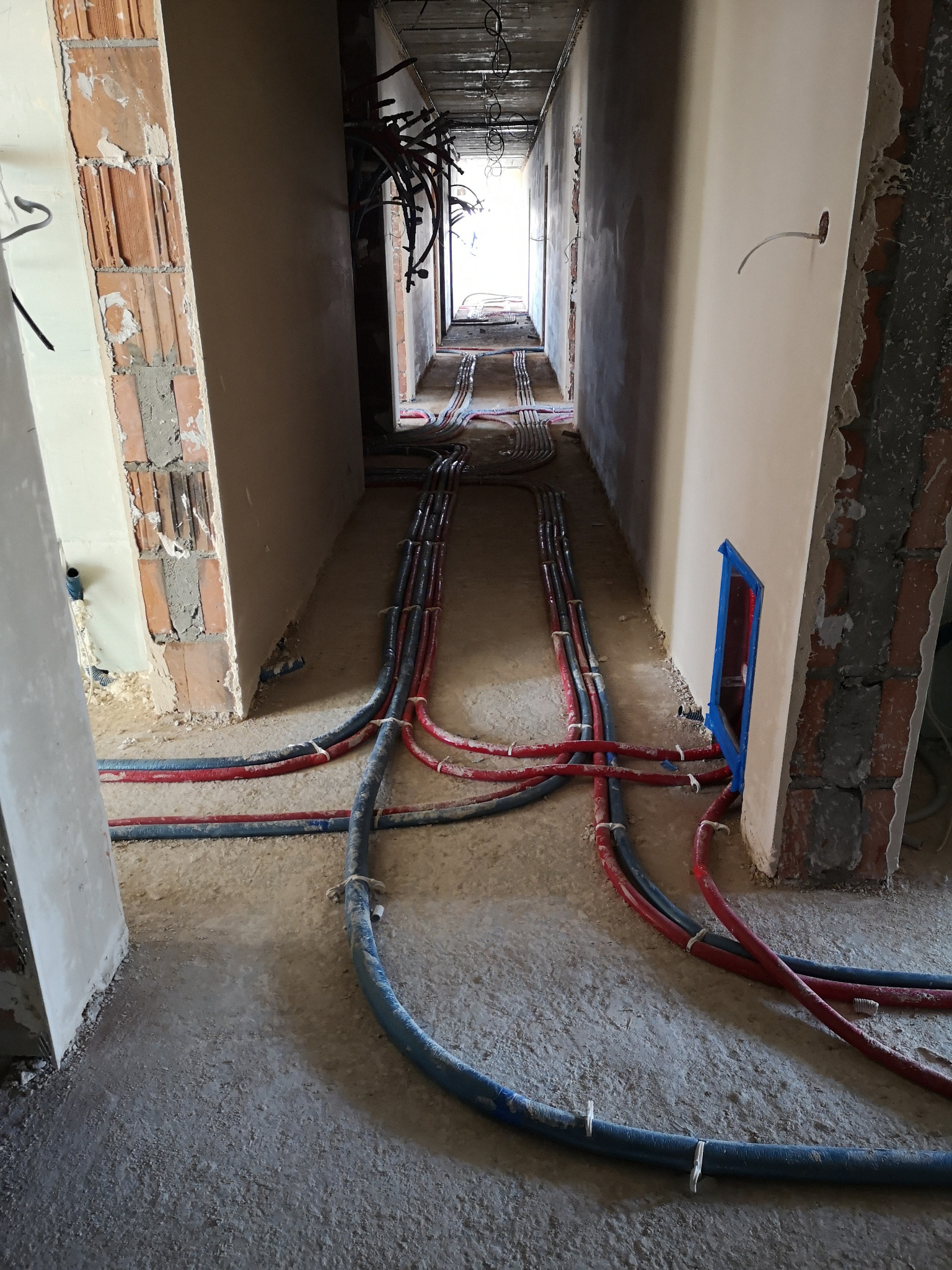 Korytarz w trakcie remontu z widocznymi ułożonymi rurami instalacji wodnej i grzewczej, mocowanymi plastikowymi opaskami do podłoża, ściany w stanie surowym.