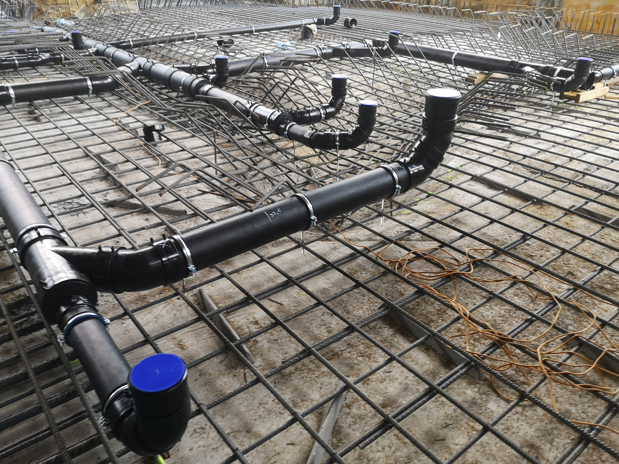 Przygotowanie instalacji kanalizacyjnej pod posadzką, widoczne czarne rury z niebieskimi zaślepkami, ułożone na siatce zbrojeniowej, w tle szalunki.