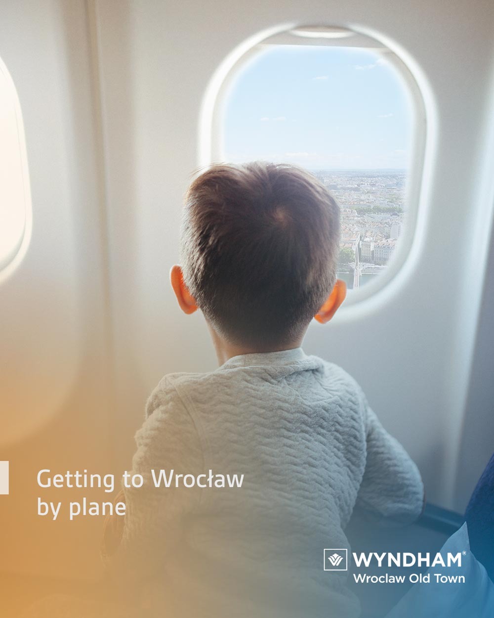 Chłopiec patrzy przez okno samolotu na panoramę Wrocławia w pogodny dzień, widoczne mosty i budynki, z tyłu głowa dziecka w szarej bluzie.