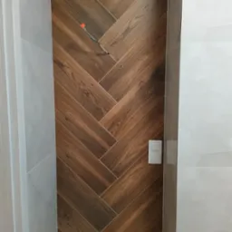 Łazienka w trakcie remontu: ściana wyłożona płytkami imitującymi drewno w jodełkę, widoczne wystające przewody i zamontowane baterie.