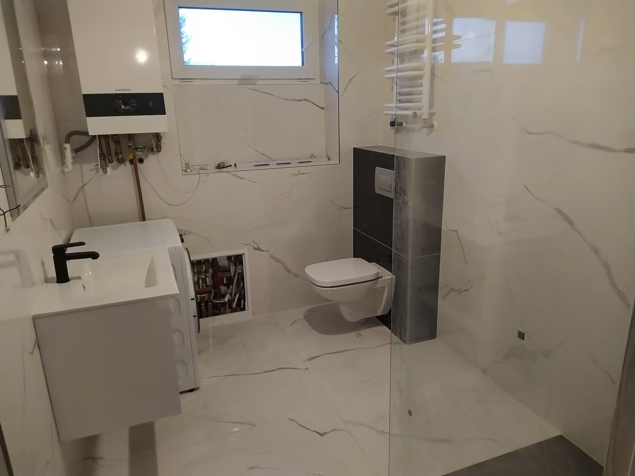 Nowoczesna łazienka z białymi marmurkowymi płytkami, wiszącą toaletą, białą umywalką z czarną baterią i grzejnikiem drabinkowym. Widoczny piec gazowy i rewizja instalacji.