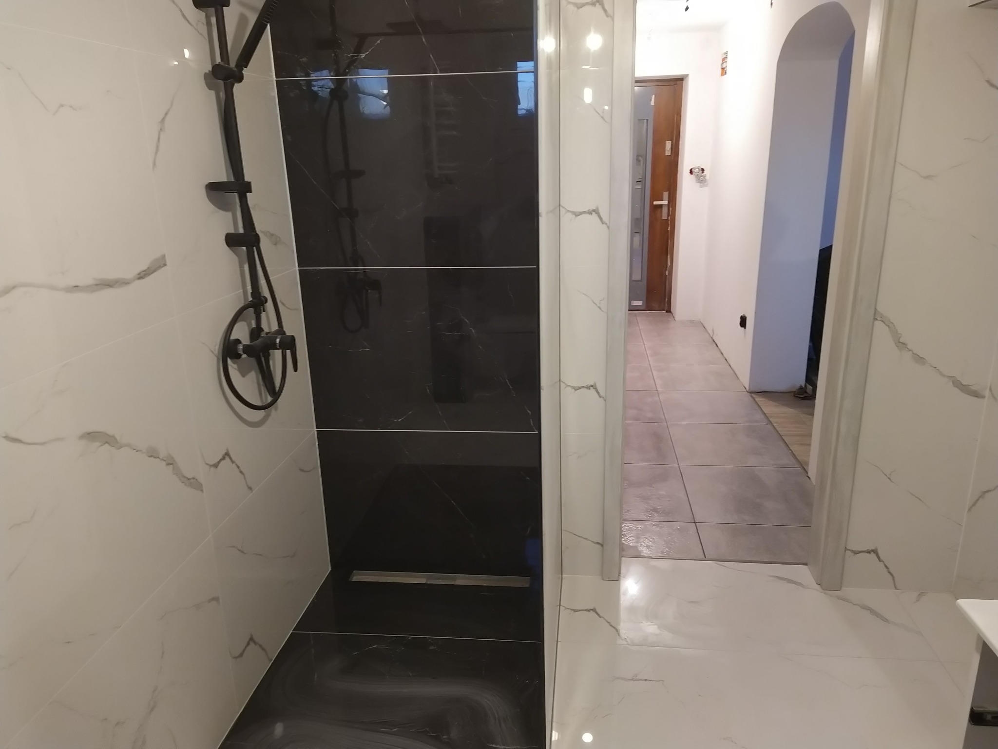 Nowoczesna łazienka z kabiną prysznicową typu walk-in, wyłożoną czarno-białymi płytkami imitującymi marmur, z czarną armaturą i odpływem liniowym, widok na wykończony korytarz z szarą podłogą.