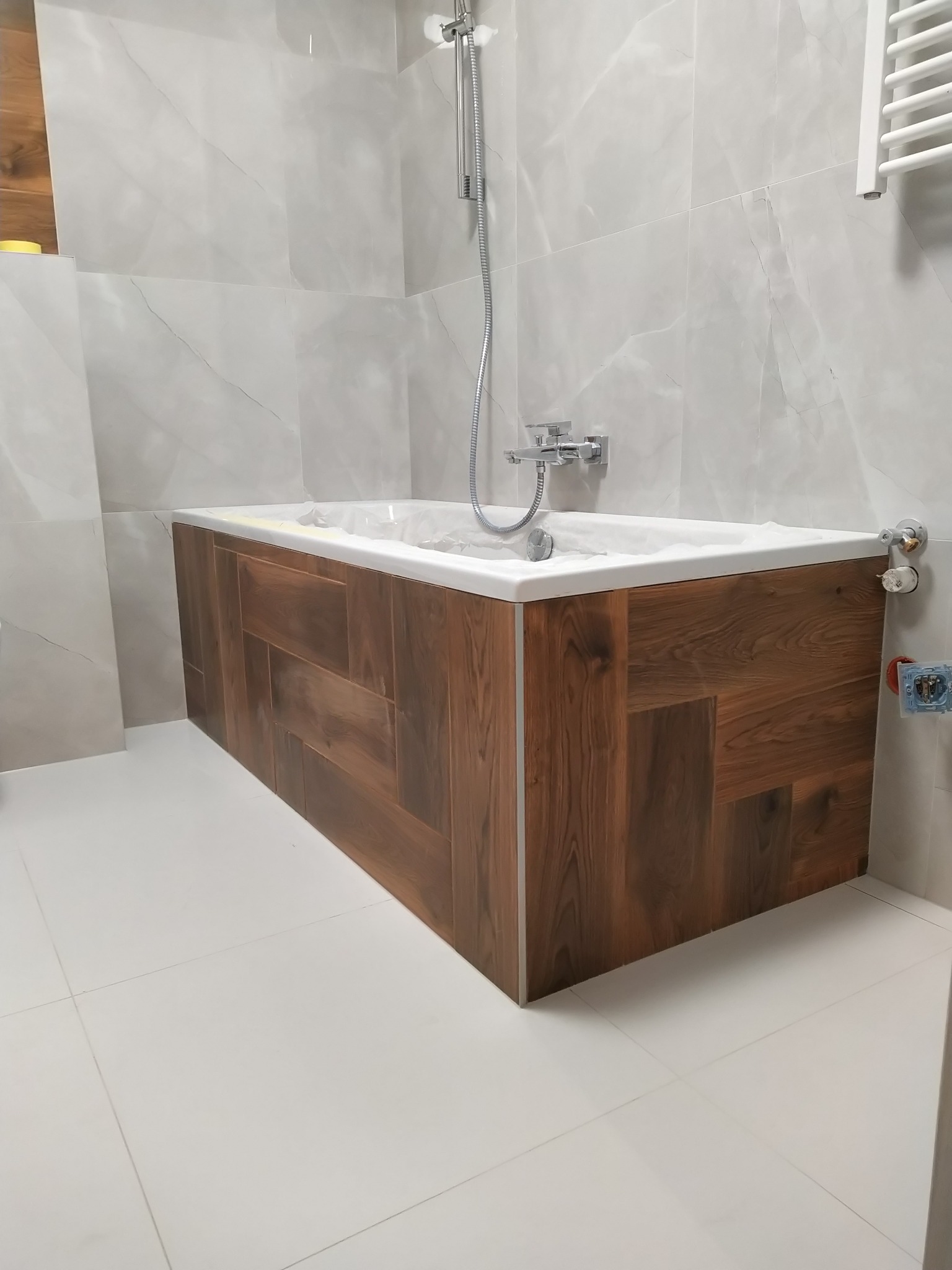 Nowoczesna łazienka z wanną obudowaną panelami imitującymi drewno, białe płytki podłogowe i ścienne, chromowana bateria i prysznic.