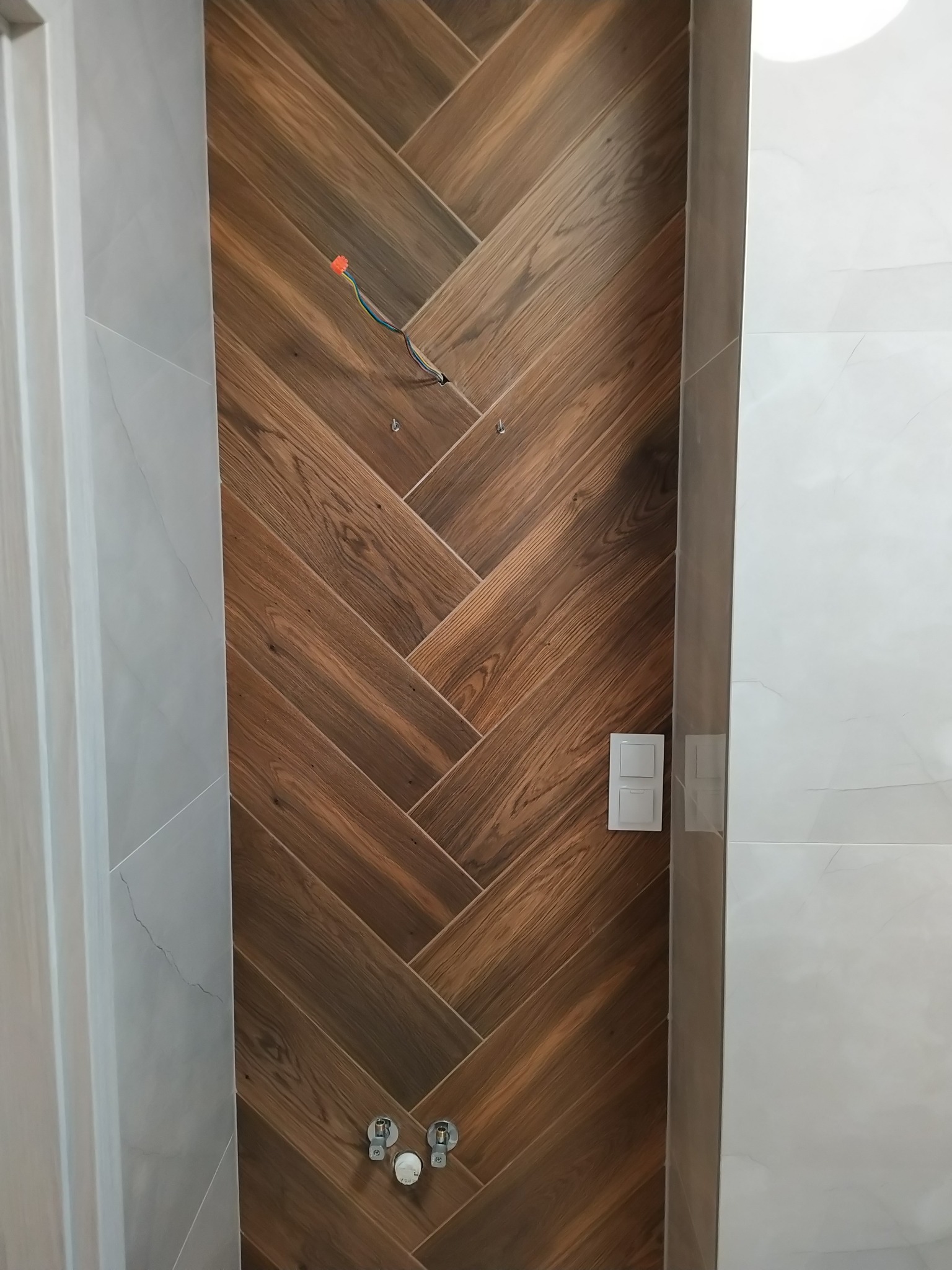 Łazienka w trakcie remontu: ściana wyłożona płytkami imitującymi drewno w jodełkę, widoczne wystające przewody i zamontowane baterie.