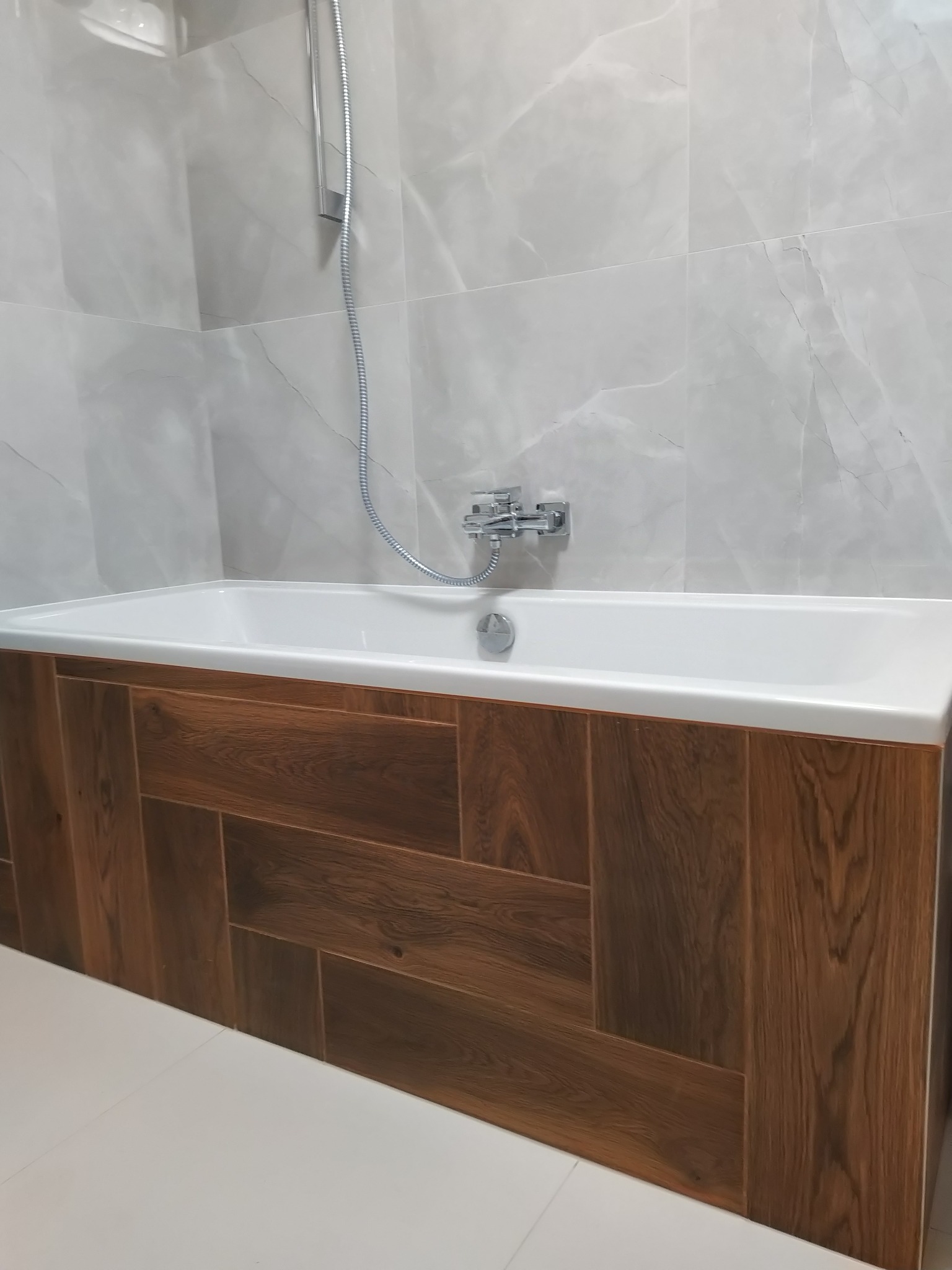 Biała wanna z obudową z paneli drewnopodobnych ułożonych w jodełkę, na tle jasnoszarych płytek imitujących marmur, bateria wannowa i słuchawka prysznicowa.