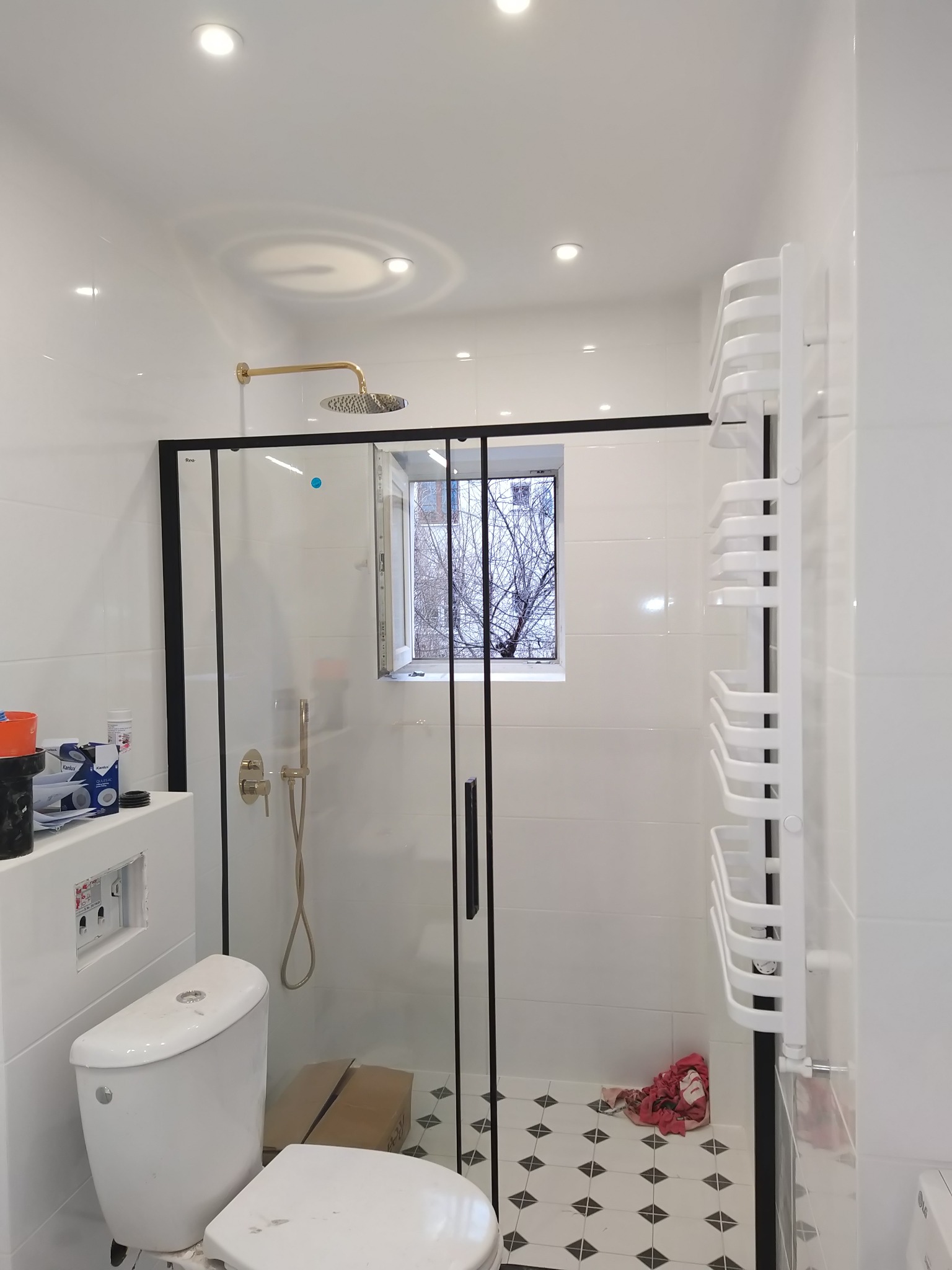 Nowoczesna łazienka z kabiną prysznicową z czarną ramą, złotą armaturą i grzejnikiem drabinkowym, widok na okno z drzewem, częściowo wykończona toaleta i karton na podłodze.