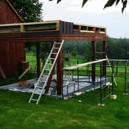 Konstrukcja z drewna w trakcie budowy, prawdopodobnie altana lub pergola, z widocznymi elementami dachu i schodami, na tle zielonego trawnika i drzew.