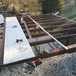 Konstrukcja z belek z częściowo zamontowanym drewnianym dachem, widoczne narzędzia pracy: wkrętarki, młotek gumowy i kompresor ze zszywaczem pneumatycznym.