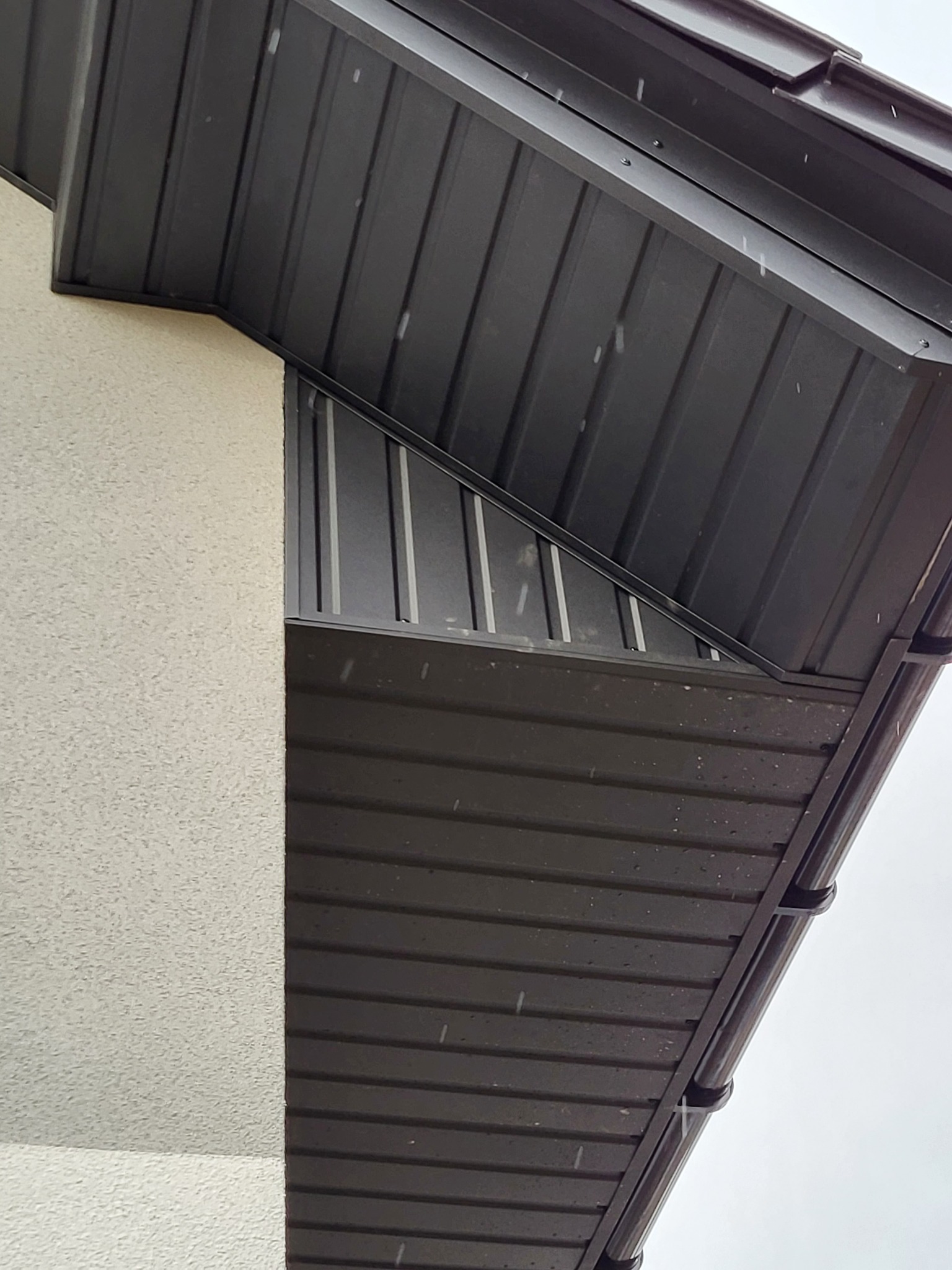 Fragment dachu z blachy trapezowej w kolorze antracytowym, widoczne mocowania rynny, elewacja budynku w jasnym kolorze z fakturą baranka, padający deszcz.