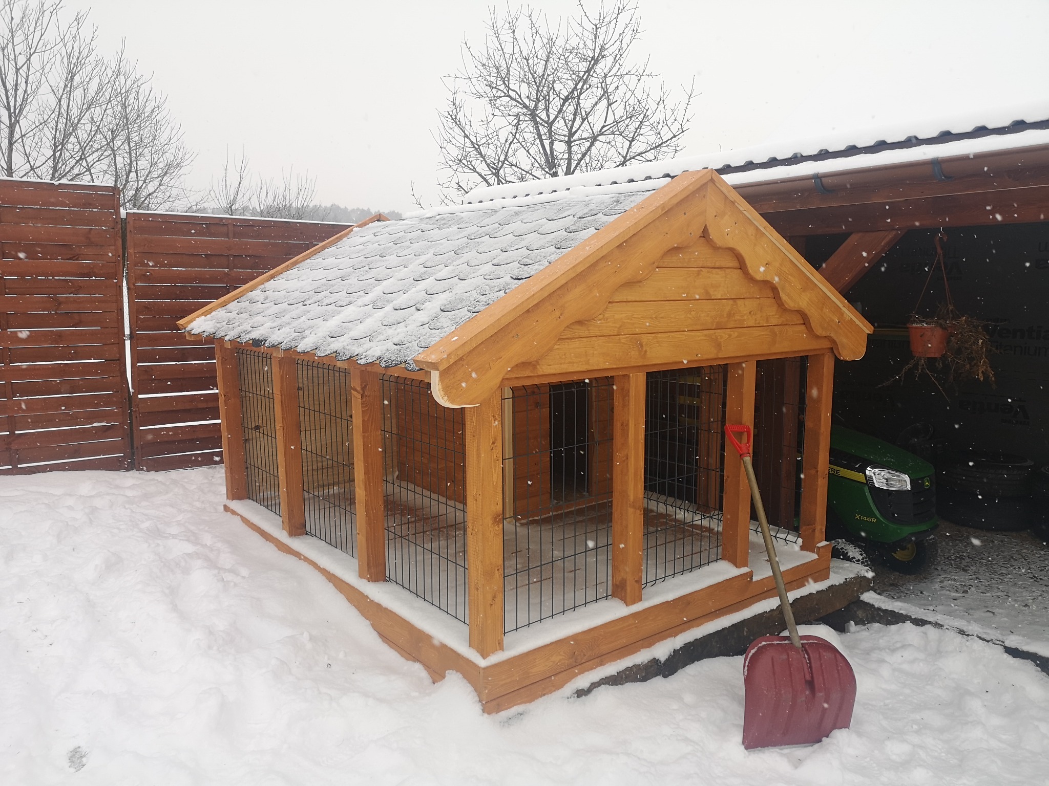 Drewniana pergola z metalowymi kratami, pokryta dachówką bitumiczną, zasypana śniegiem, z czerwoną łopatą opartą o bok, na tle ogrodzenia i traktorka.