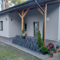 Szary dom jednorodzinny w Bądkowie po malowaniu elewacji, z drewnianą konstrukcją werandy i dekoracyjnym murkiem z betonowych pustaków przed wejściem.