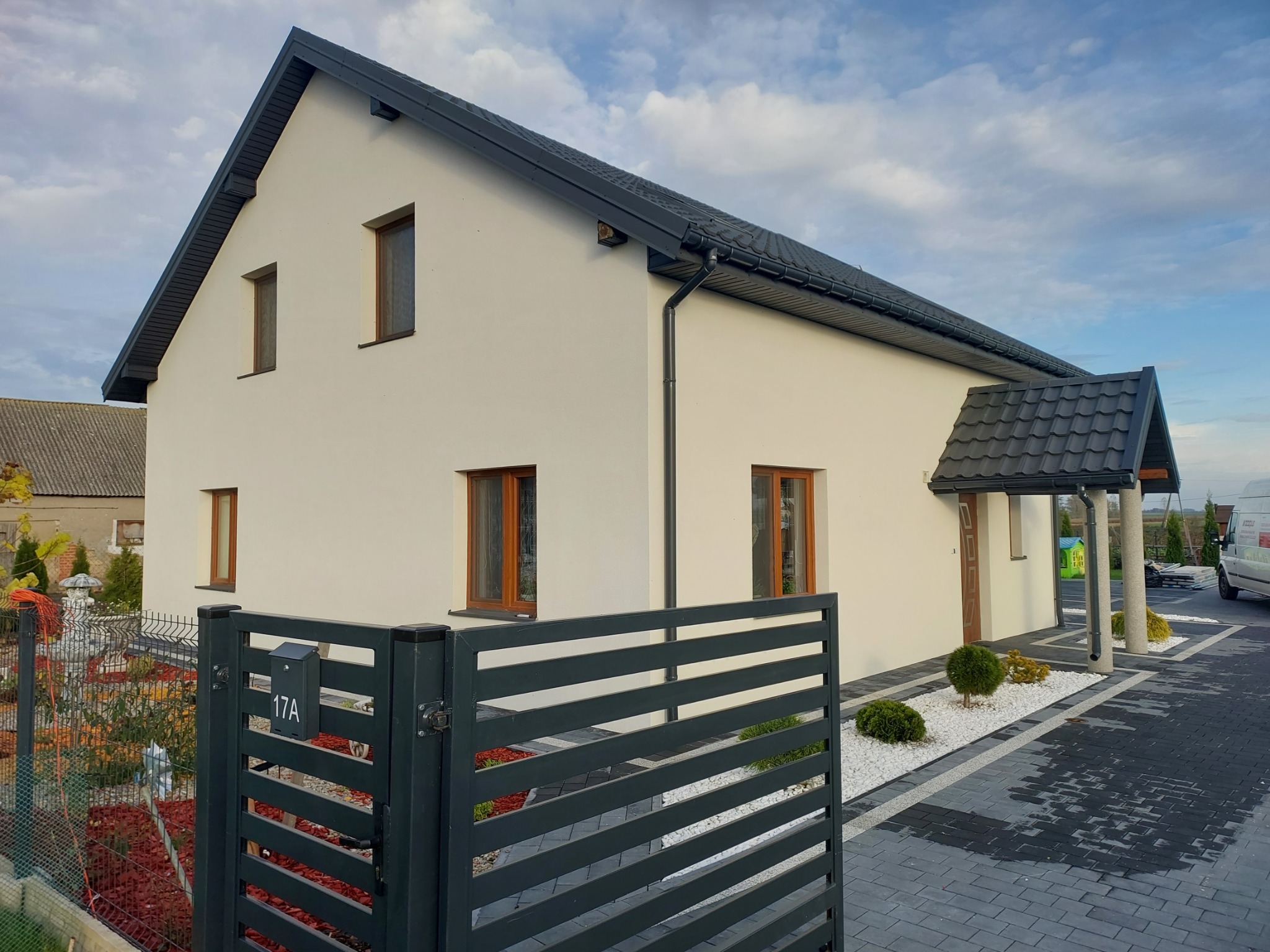 Dwukondygnacyjny dom z jasną elewacją, ciemną dachówką i stolarką okienną, widoczny zza grafitowego ogrodzenia z numerem 17A, z zadbanym otoczeniem
