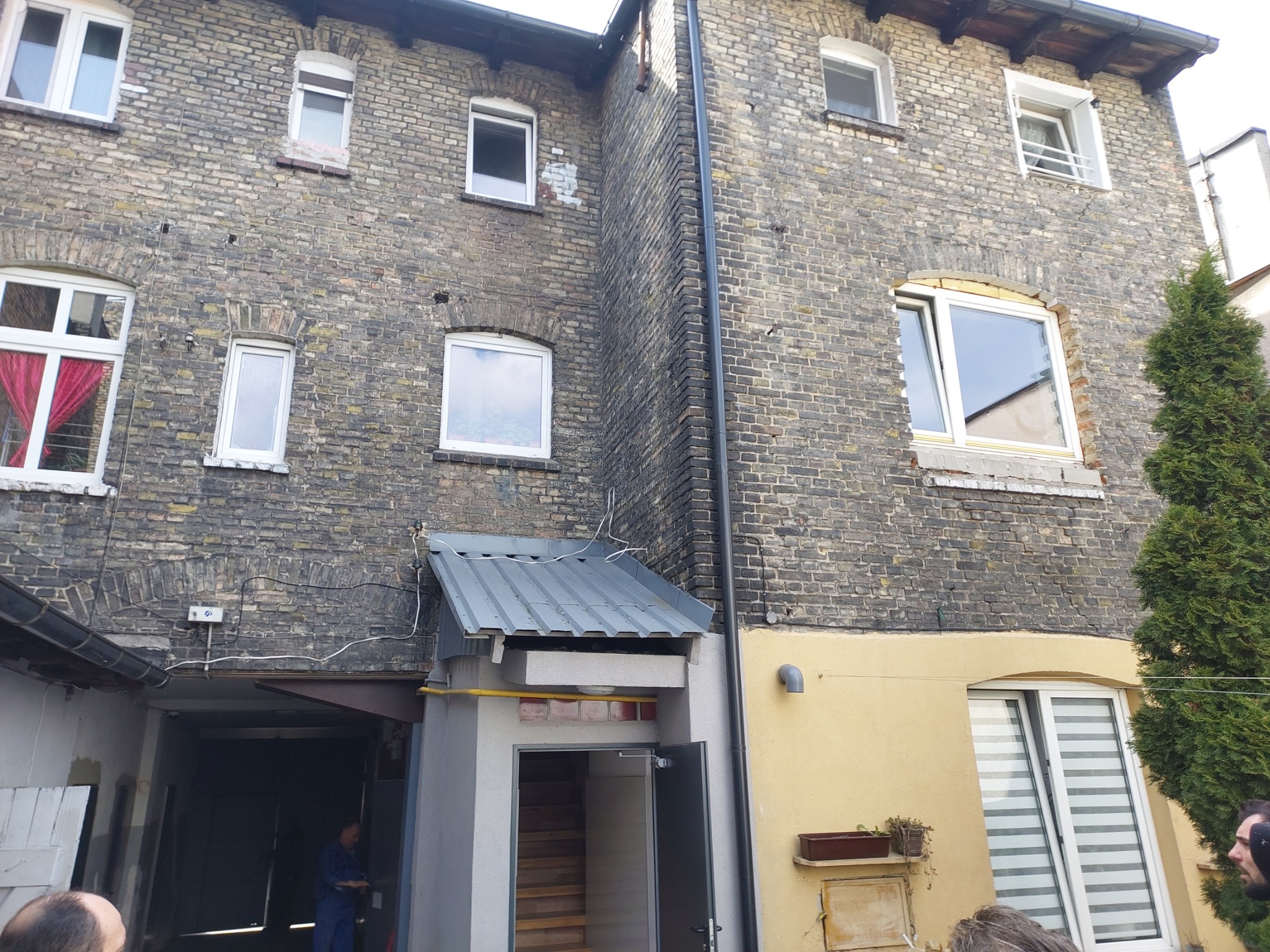 Dwukondygnacyjny budynek z cegły z oknami, zadaszonym wejściem i fragmentem żółtej elewacji po prawej stronie, widoczne ślady zabrudzeń na cegłach.