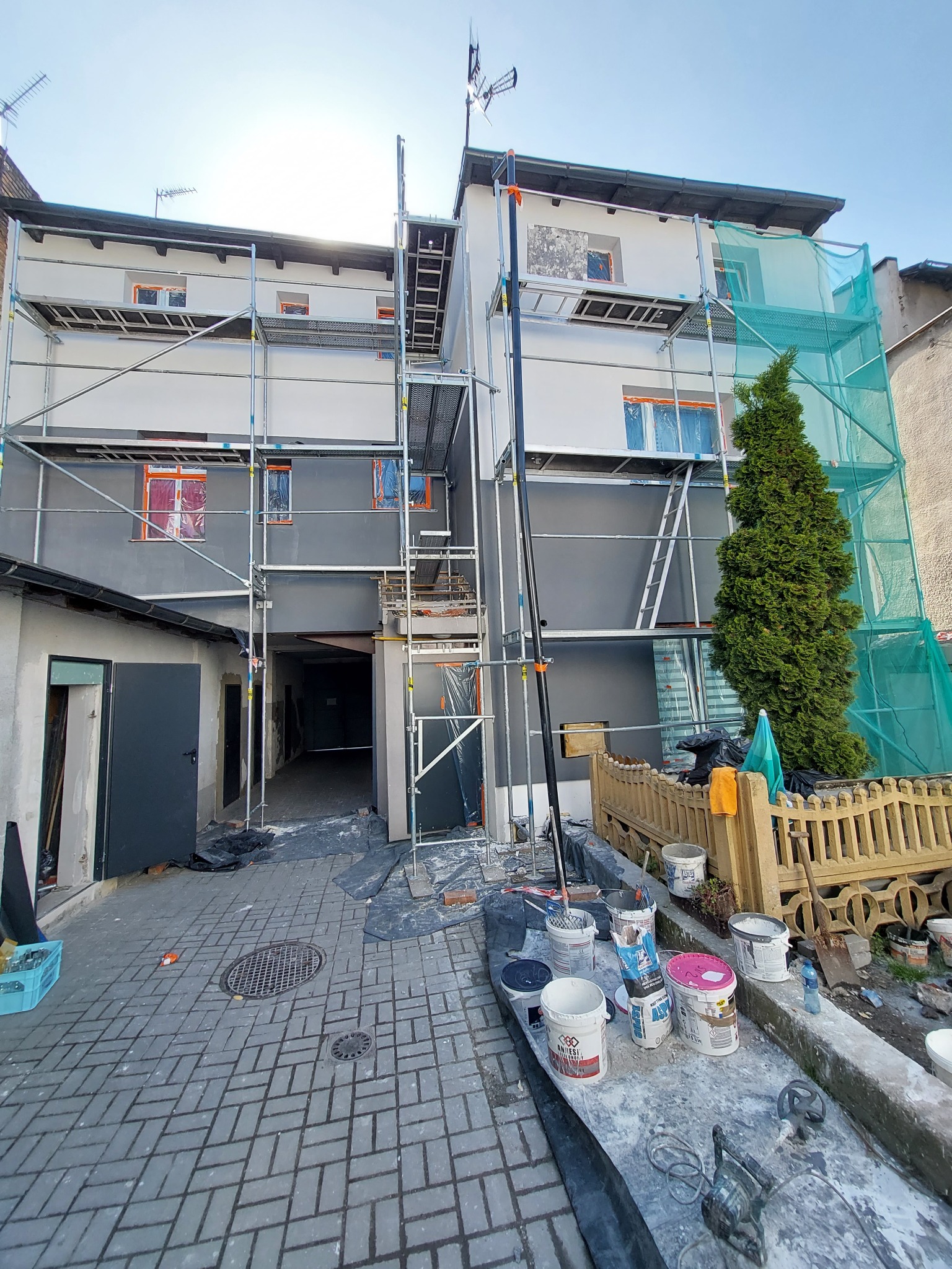 Budynek w trakcie malowania elewacji, widoczne rusztowania, zabezpieczone okna folią, oraz wiaderka z farbą i narzędzia malarskie na pierwszym planie.