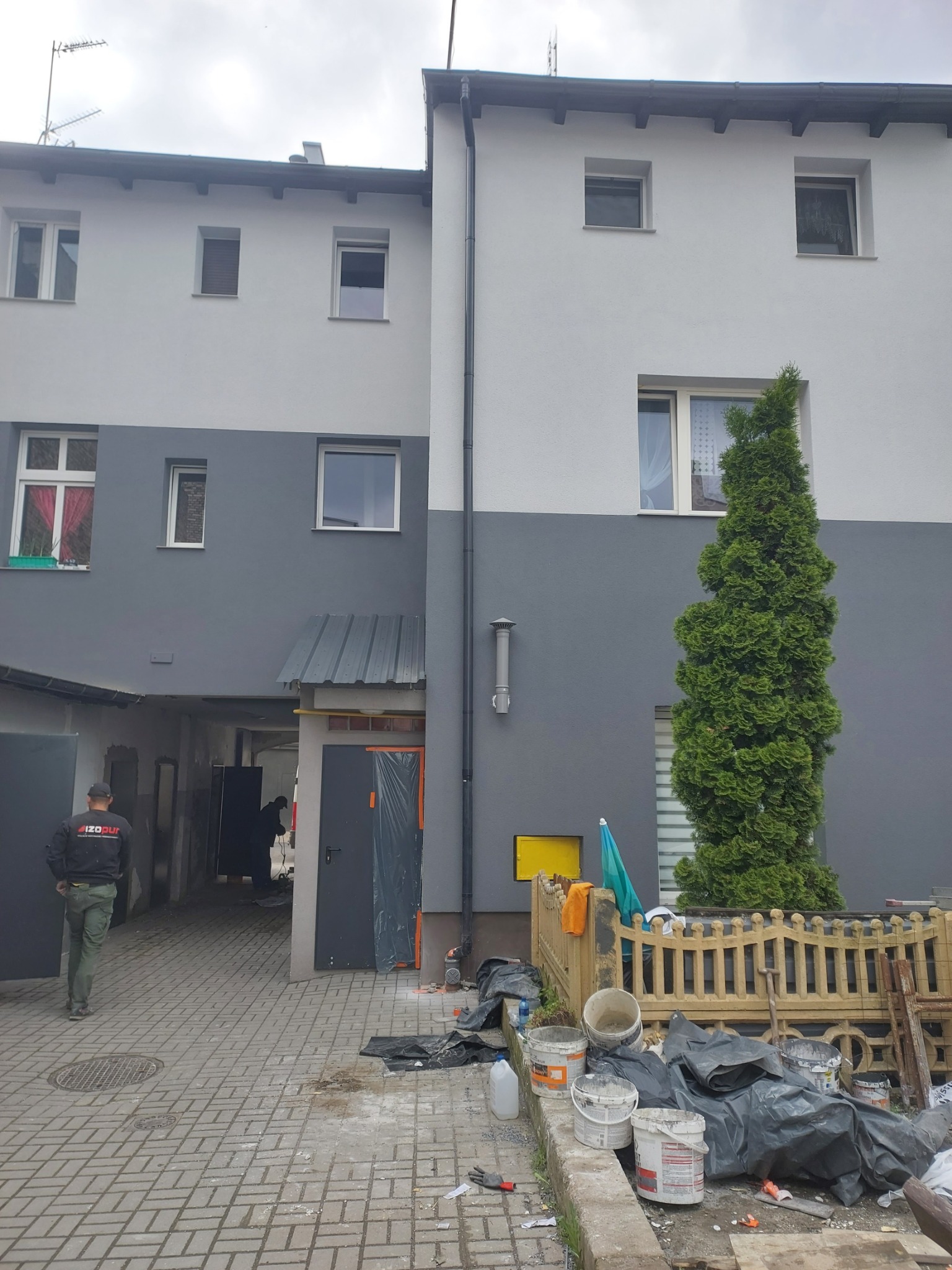 Dwukolorowa fasada budynku po malowaniu, widoczne ślady prac wykończeniowych, narzędzia i materiały budowlane na pierwszym planie. Pracownik w uniformie w oddali.