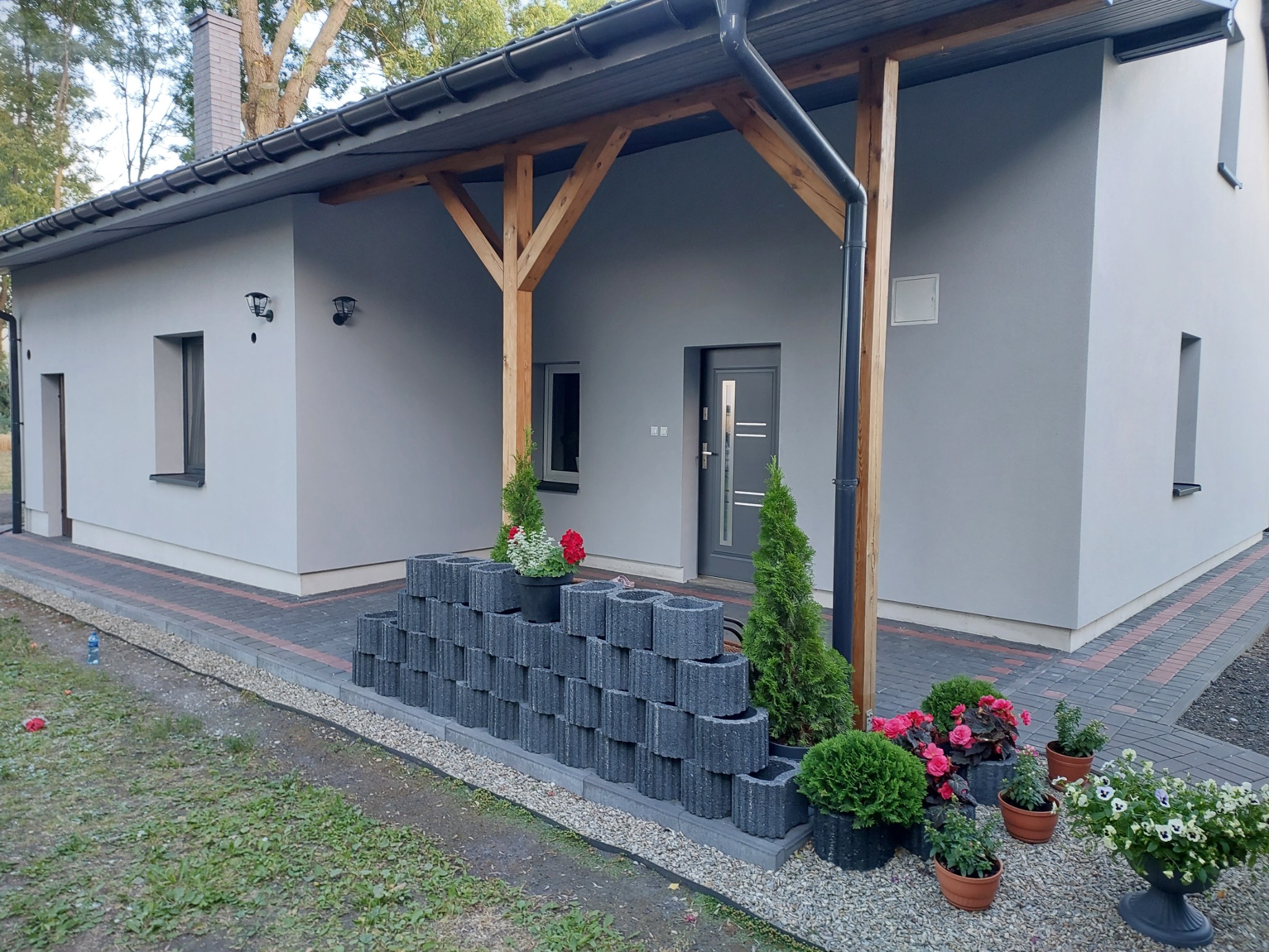 Szary dom jednorodzinny w Bądkowie po malowaniu elewacji, z drewnianą konstrukcją werandy i dekoracyjnym murkiem z betonowych pustaków przed wejściem.