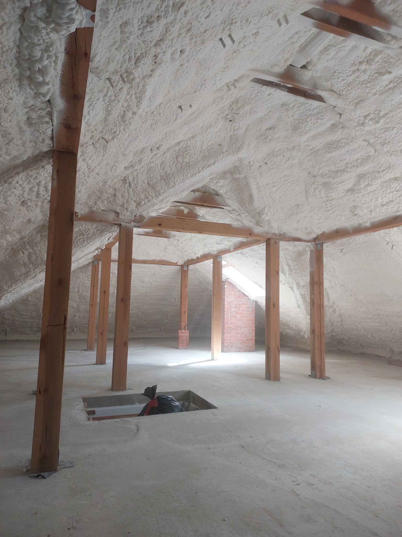 Poddasze z drewnianą konstrukcją po ociepleniu pianką PUR; widoczne nierównomierne pokrycie pianką i otwór w podłodze z workiem gruzu.