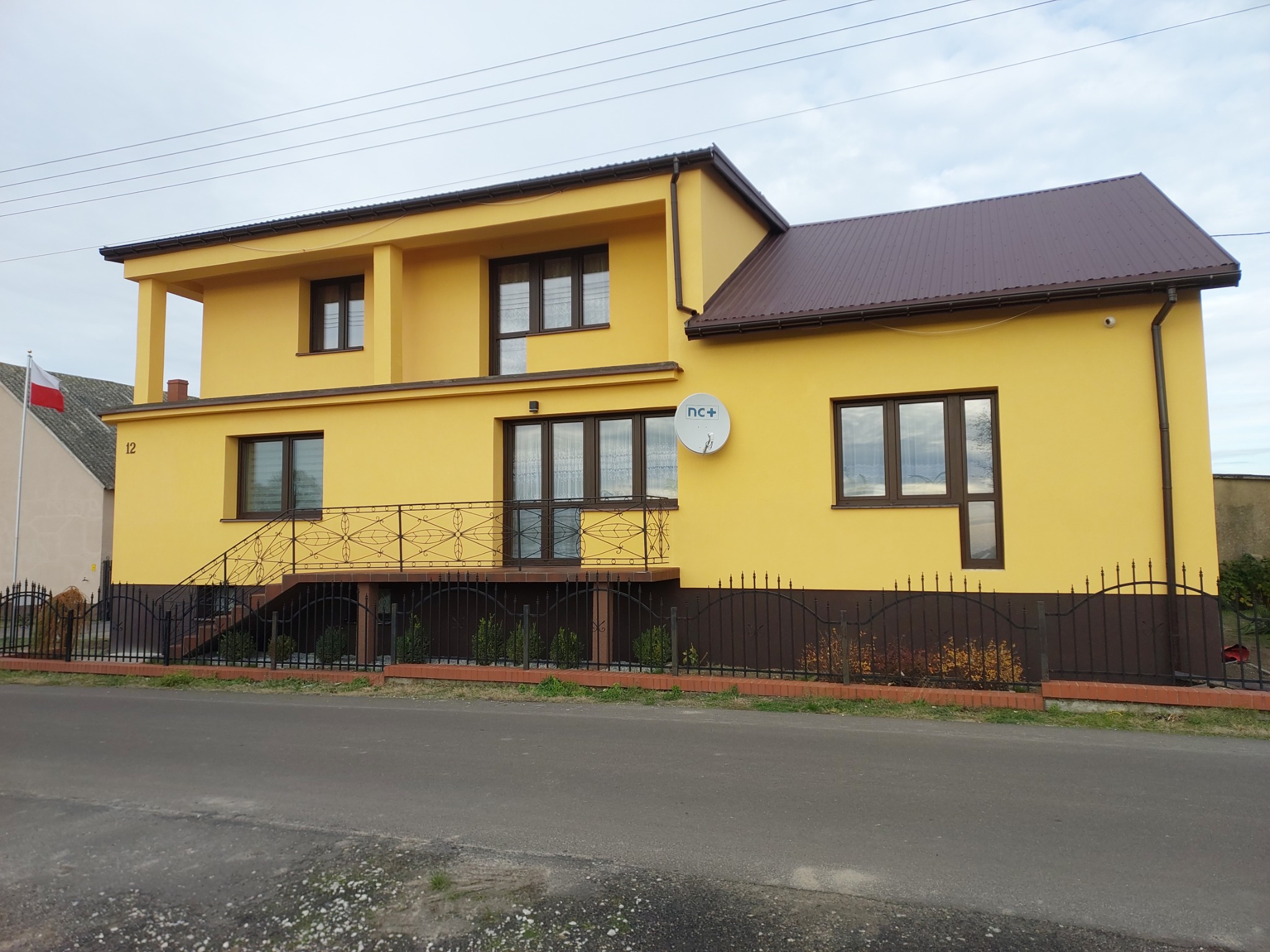 Dwupiętrowy dom w odcieniach żółci z brązowymi akcentami, widok z ulicy, z widoczną anteną satelitarną i fragmentem polskiej flagi w tle.
