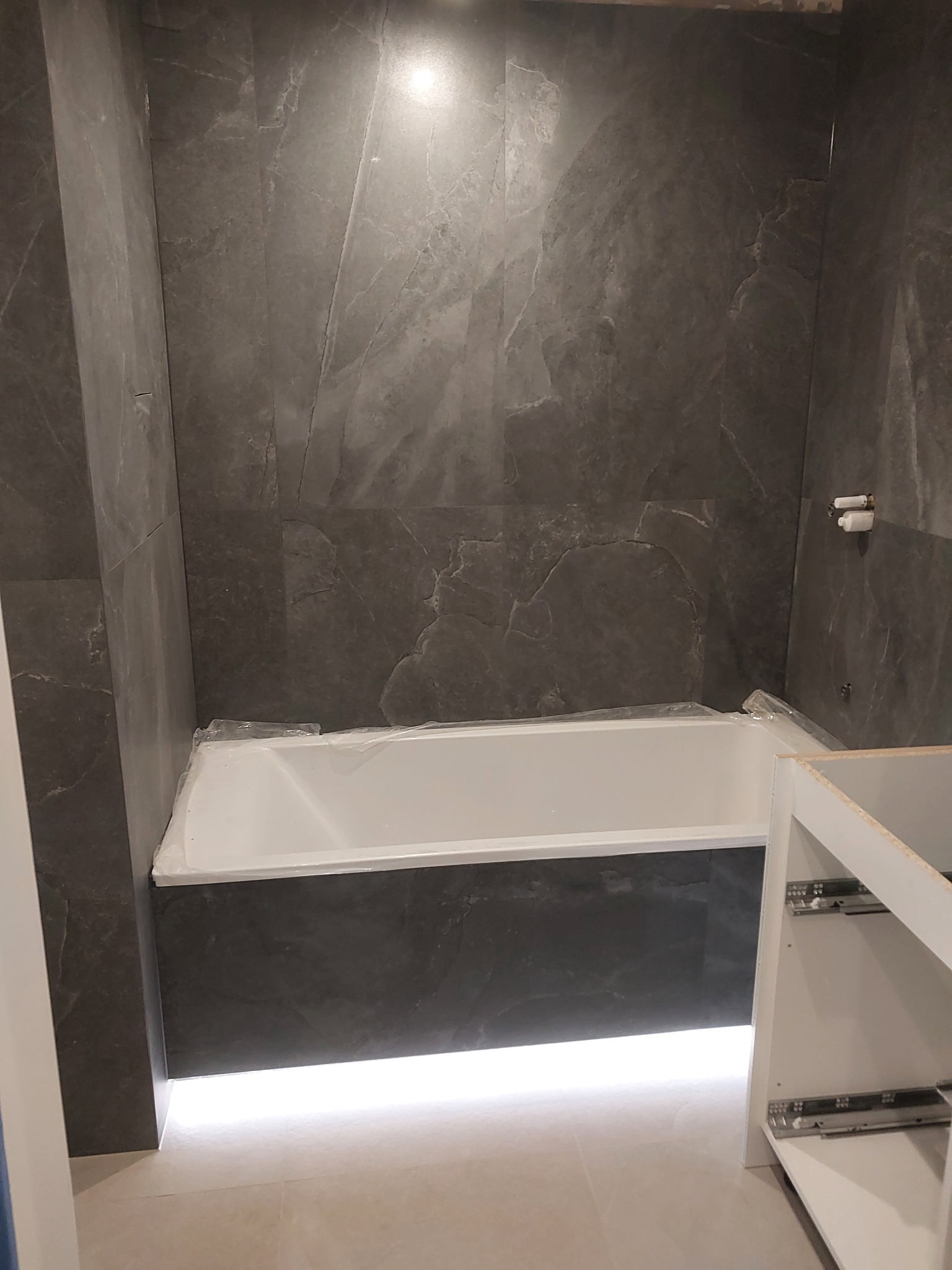 Montaż wanny z szarą okładziną imitującą marmur, widoczne fragmenty białych mebli łazienkowych i podświetlenie LED pod wanną, zabezpieczona folią ochronną.