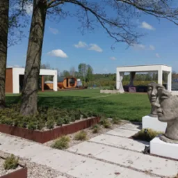 Nowoczesna aranżacja ogrodu z drewnianą sauną beczką, jacuzzi w zabudowie, altaną z zasłonami i rzeźbą dwóch twarzy na tle zielonego trawnika i błękitnego nieba.