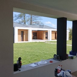 Mariusz Skora - Widok z zadaszonego tarasu na nowoczesny dom z drewnianą elewacją i zielonym trawnikiem; na pierwszym planie narzędzia i materiały budowlane.