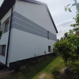 Dwukondygnacyjny dom z białą elewacją, ozdobiony trzema szarymi pasami i ciemnoszarą podmurówką, widoczny z perspektywy ogrodu w słoneczny dzień.