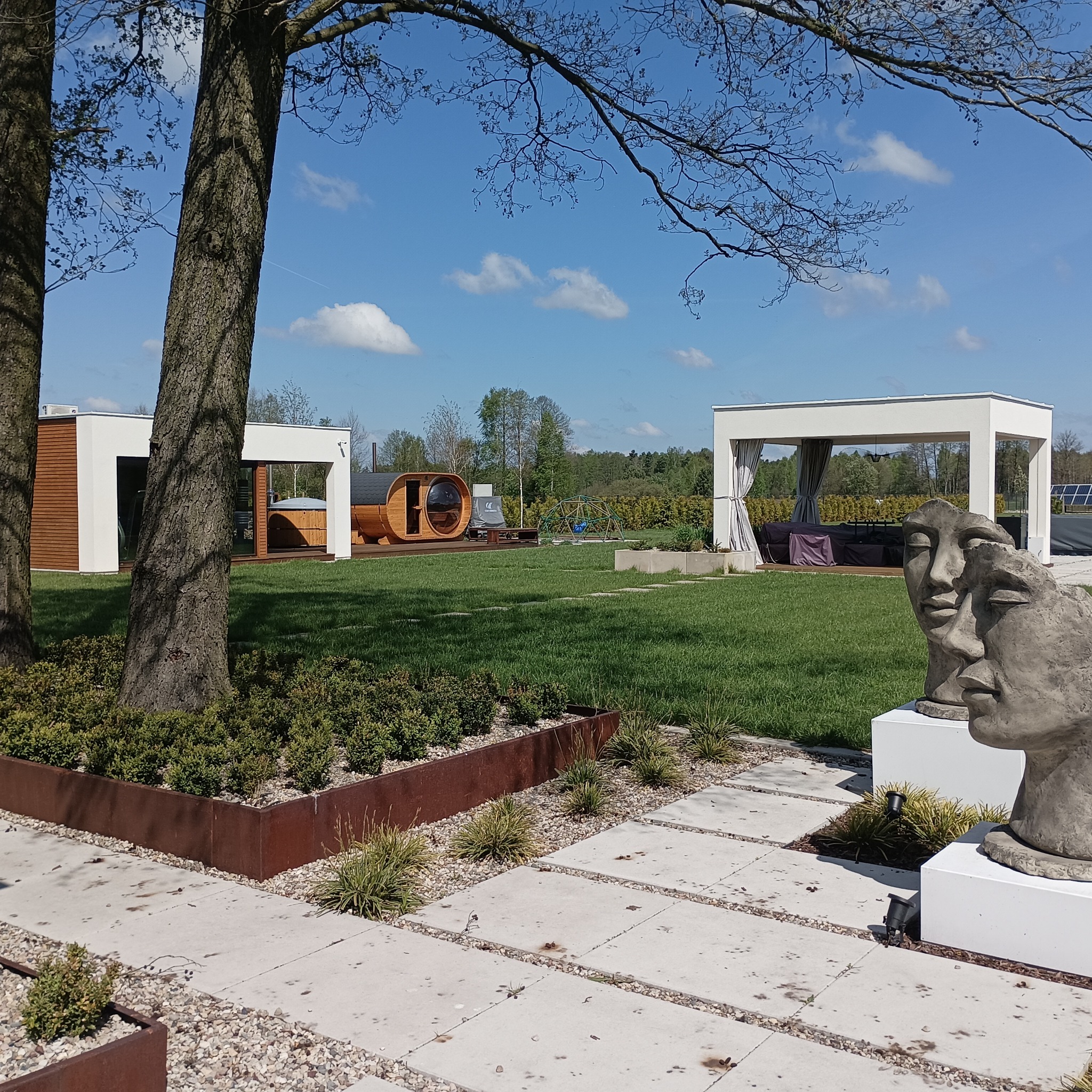 Nowoczesna aranżacja ogrodu z drewnianą sauną beczką, jacuzzi w zabudowie, altaną z zasłonami i rzeźbą dwóch twarzy na tle zielonego trawnika i błękitnego nieba.