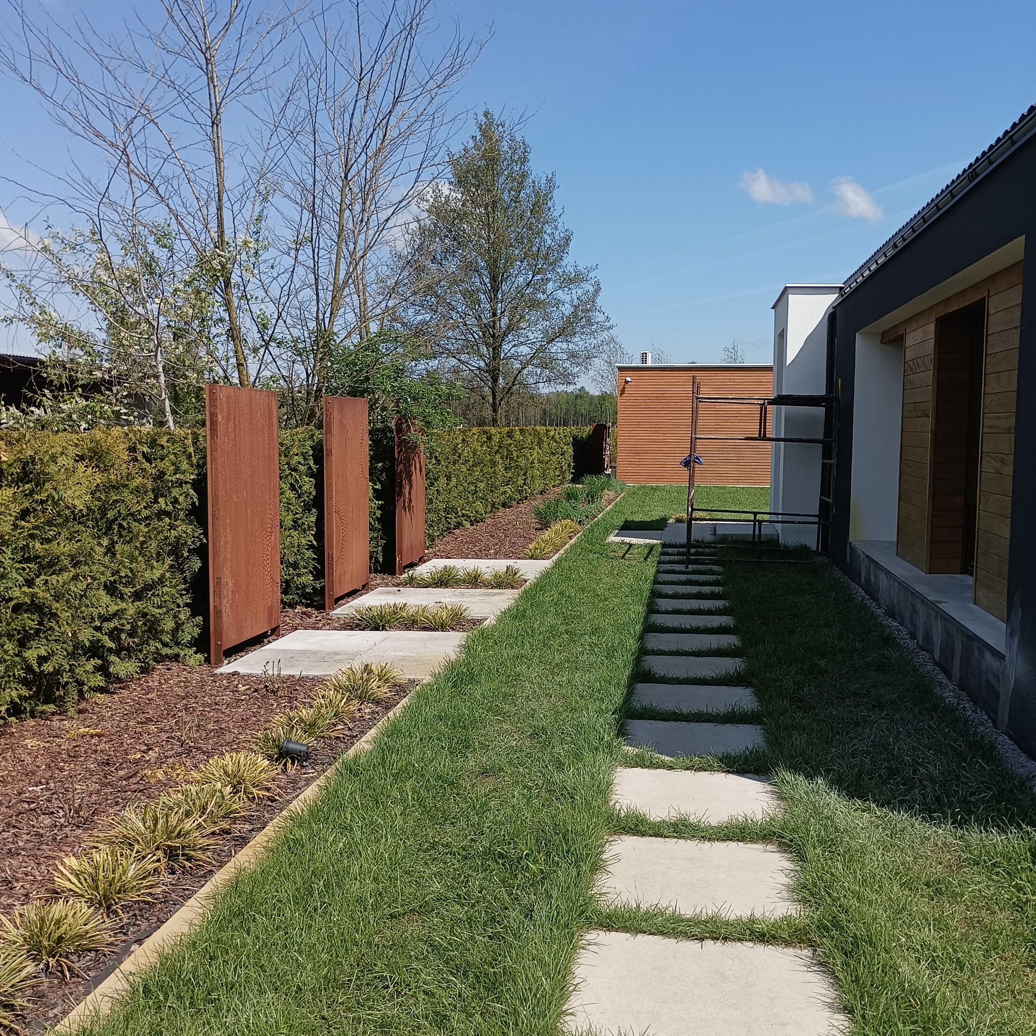 Nowoczesny ogród z betonowymi płytami chodnikowymi w trawie, rustykalnymi panelami ogrodowymi i starannie przyciętym żywopłotem.