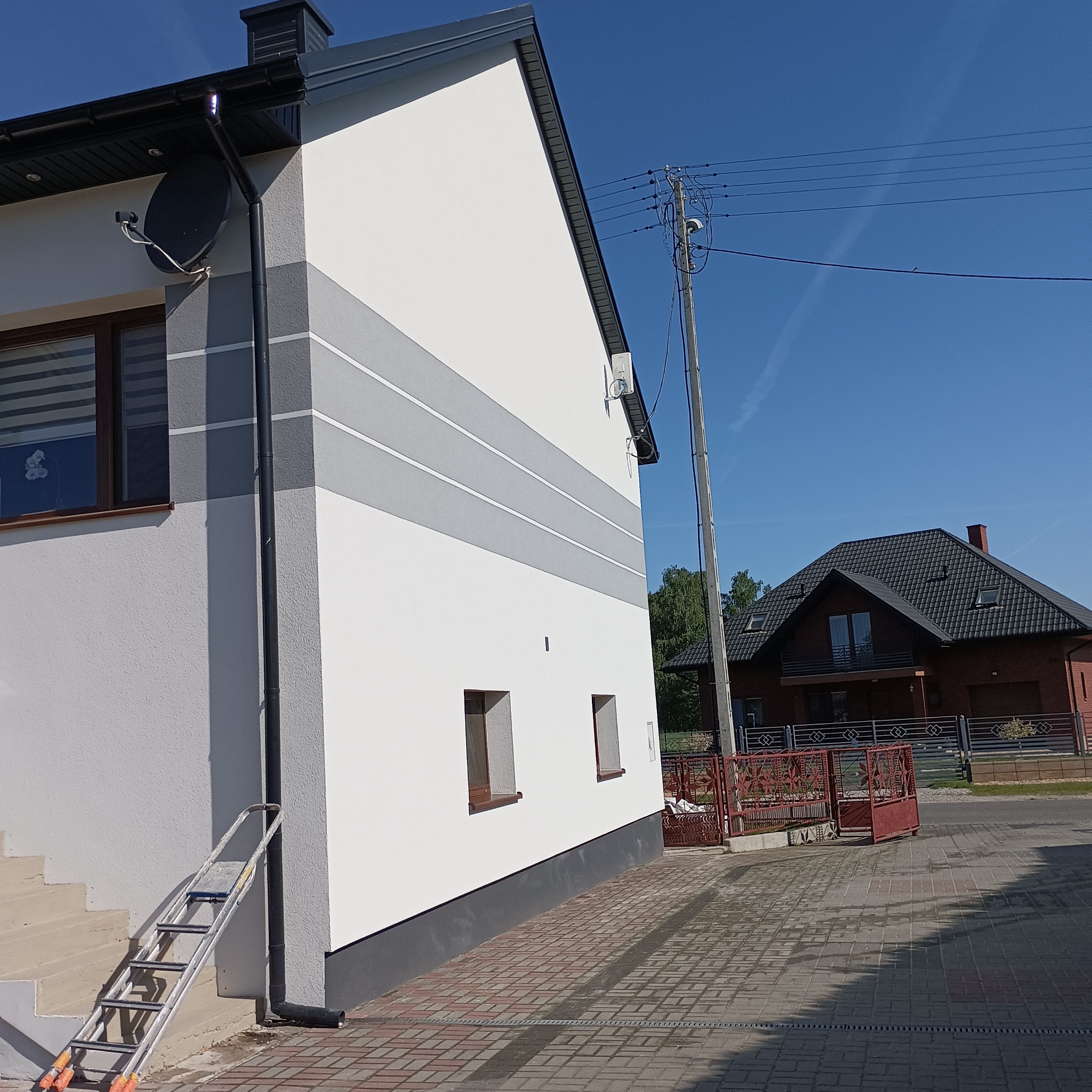 Świeżo pomalowana elewacja domu z szarymi pasami, widoczna drabina oparta o ścianę, na tle błękitne niebo i sąsiedni dom z ciemną dachówką.