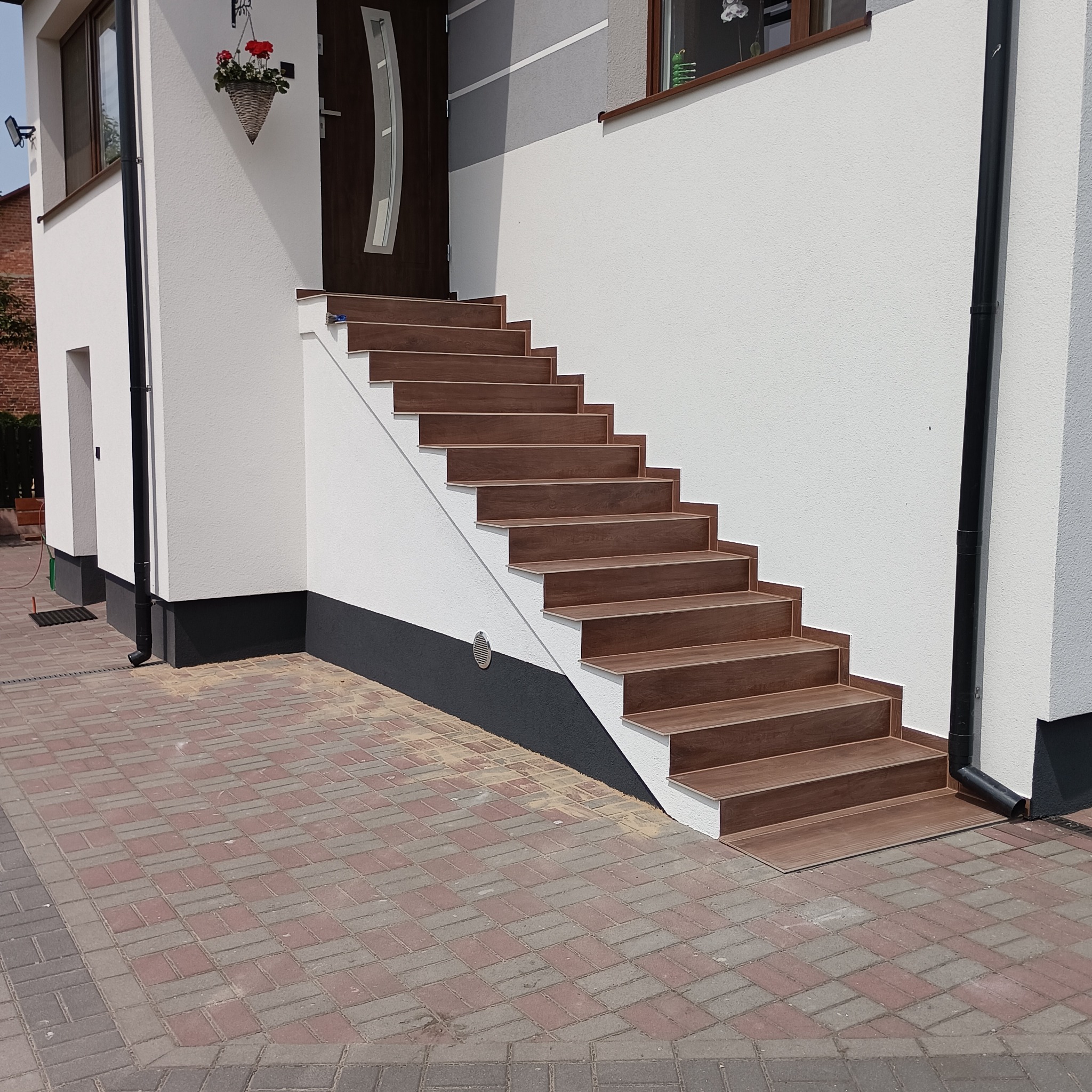 Zewnętrzne schody wejściowe do domu wyłożone brązowymi płytkami imitującymi drewno, kontrastujące z białą elewacją budynku i cokołem w kolorze grafitowym, widoczne elementy systemu rynnowego.