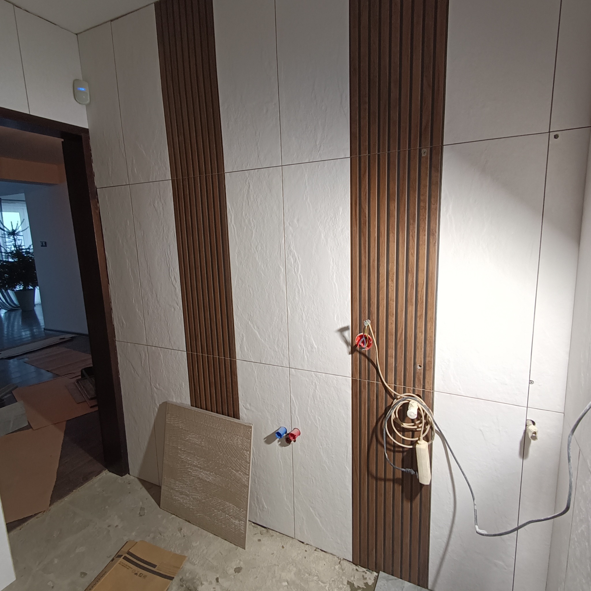 Ściana w trakcie wykańczania z białymi płytkami imitującymi kamień oraz panelami dekoracyjnymi z drewnopodobnymi lamelami, widoczne wystające kable elektryczne, rurki instalacyjne i kartony...