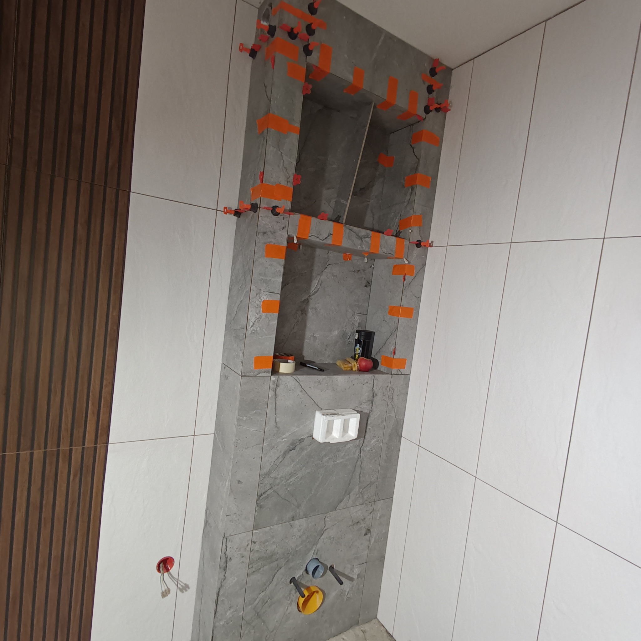 Łazienka w trakcie wykańczania: białe płytki na ścianach, szara wnęka z półkami wyłożona płytkami imitującymi kamień, pomarańczowe klipsy poziomujące płytki, widoczne przyłącza hydrauliczne...