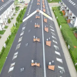Widok z góry na szereg identycznych domów jednorodzinnych z ciemnymi dachami, ceglanymi kominami i oknami dachowymi, otoczonych zielenią.