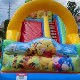 Wielobarwny dmuchany zamek ze zjeżdżalnią, ozdobiony postaciami z bajki o Kubusiu Puchatku, ustawiony na zewnątrz, widoczne drzewa i słupy energetyczne w tle.