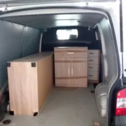 Meble z jasnego drewna zabezpieczone w przestrzeni ładunkowej białego busa z otwartą klapą bagażnika, gotowe do transportu.