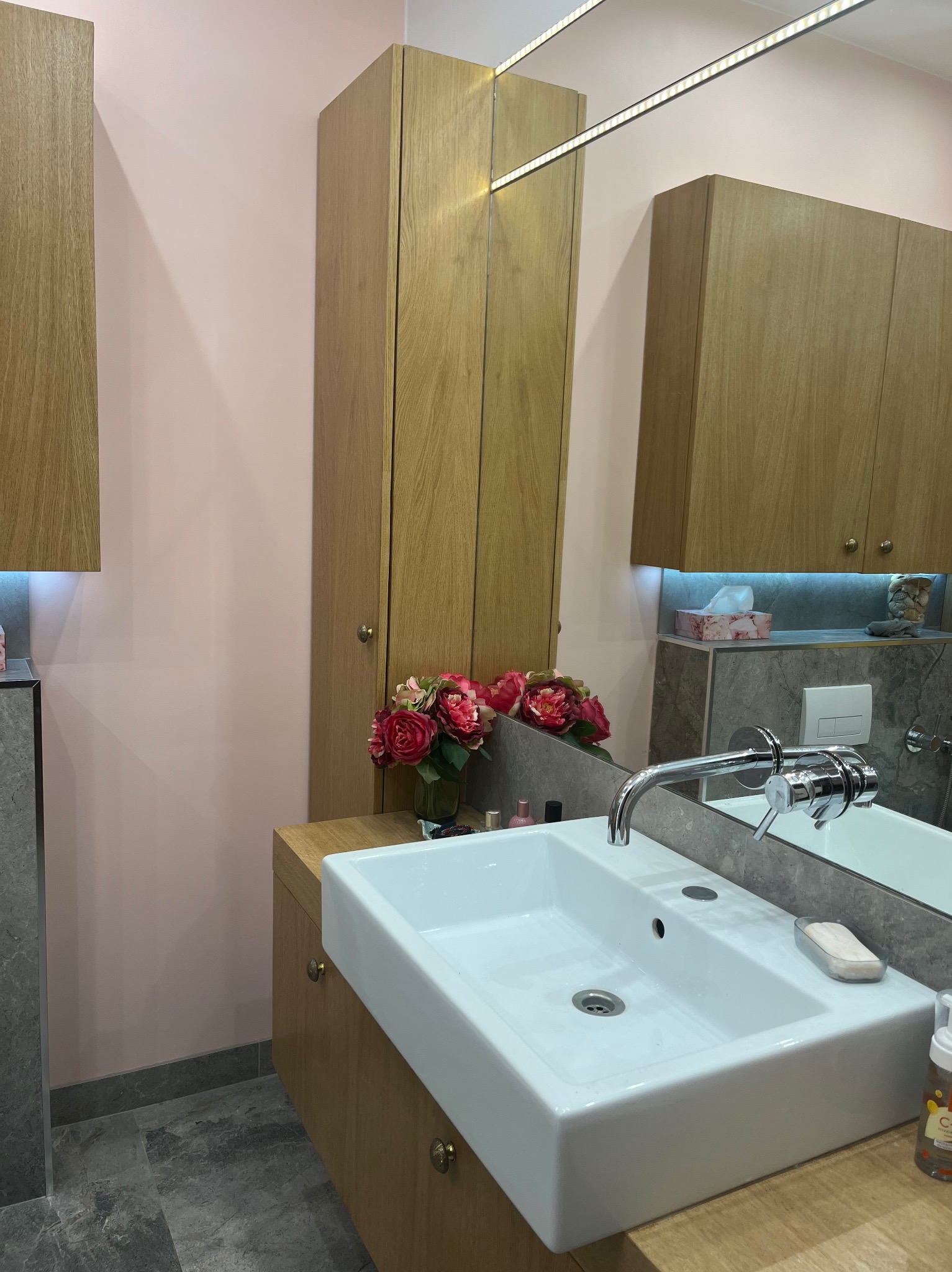 Nowoczesna łazienka z różowymi ścianami, drewnianymi szafkami, prostokątną umywalką i chromowaną baterią, z lustrem z oświetleniem LED i bukietem róż w tle.