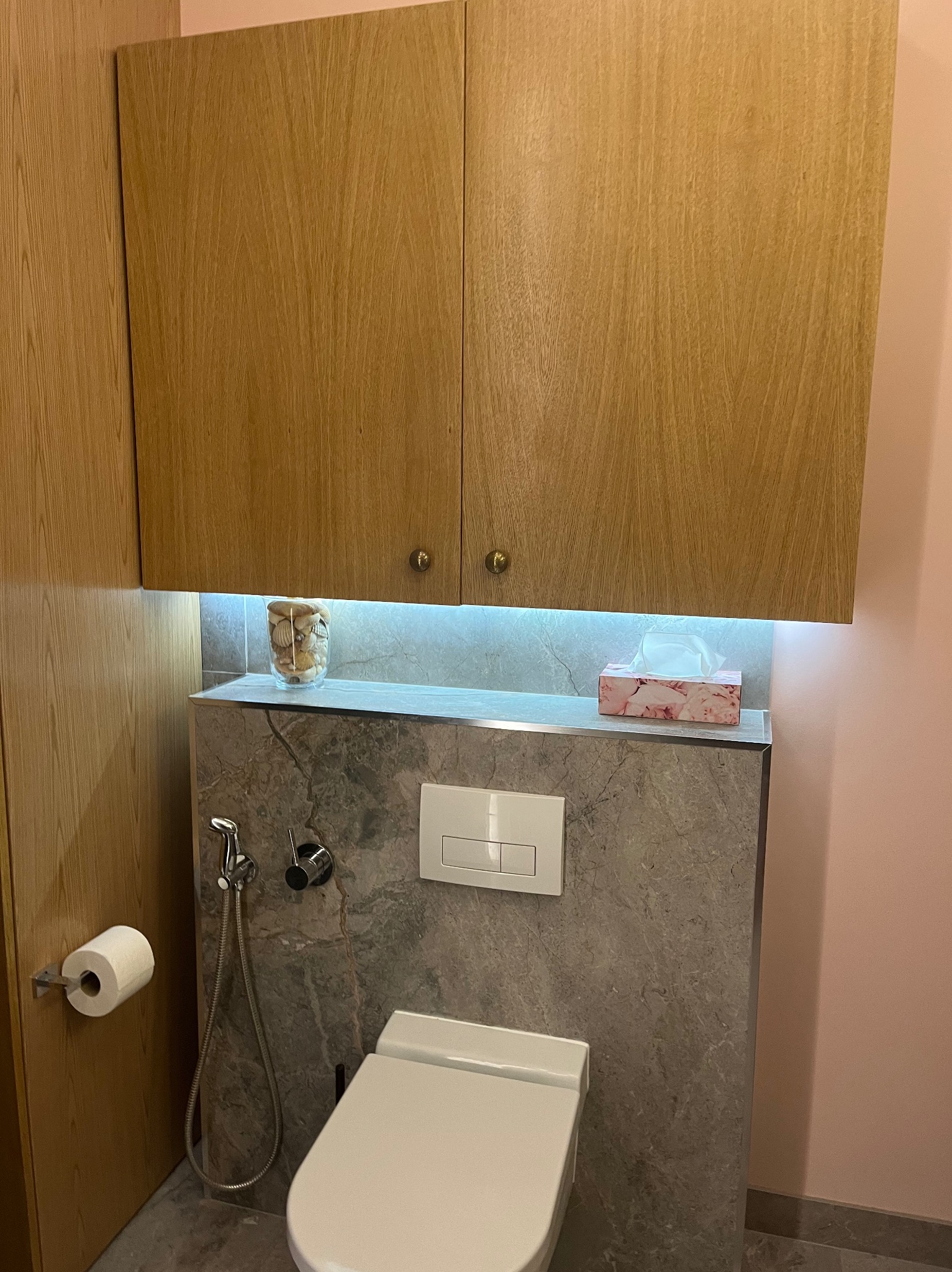 Nowoczesna toaleta: podwieszana miska WC z szarą okładziną imitującą marmur, podświetlana półka z dekoracyjnym słoikiem z muszelkami i pudełkiem chusteczek, drewniane szafki wiszące, bidetka...