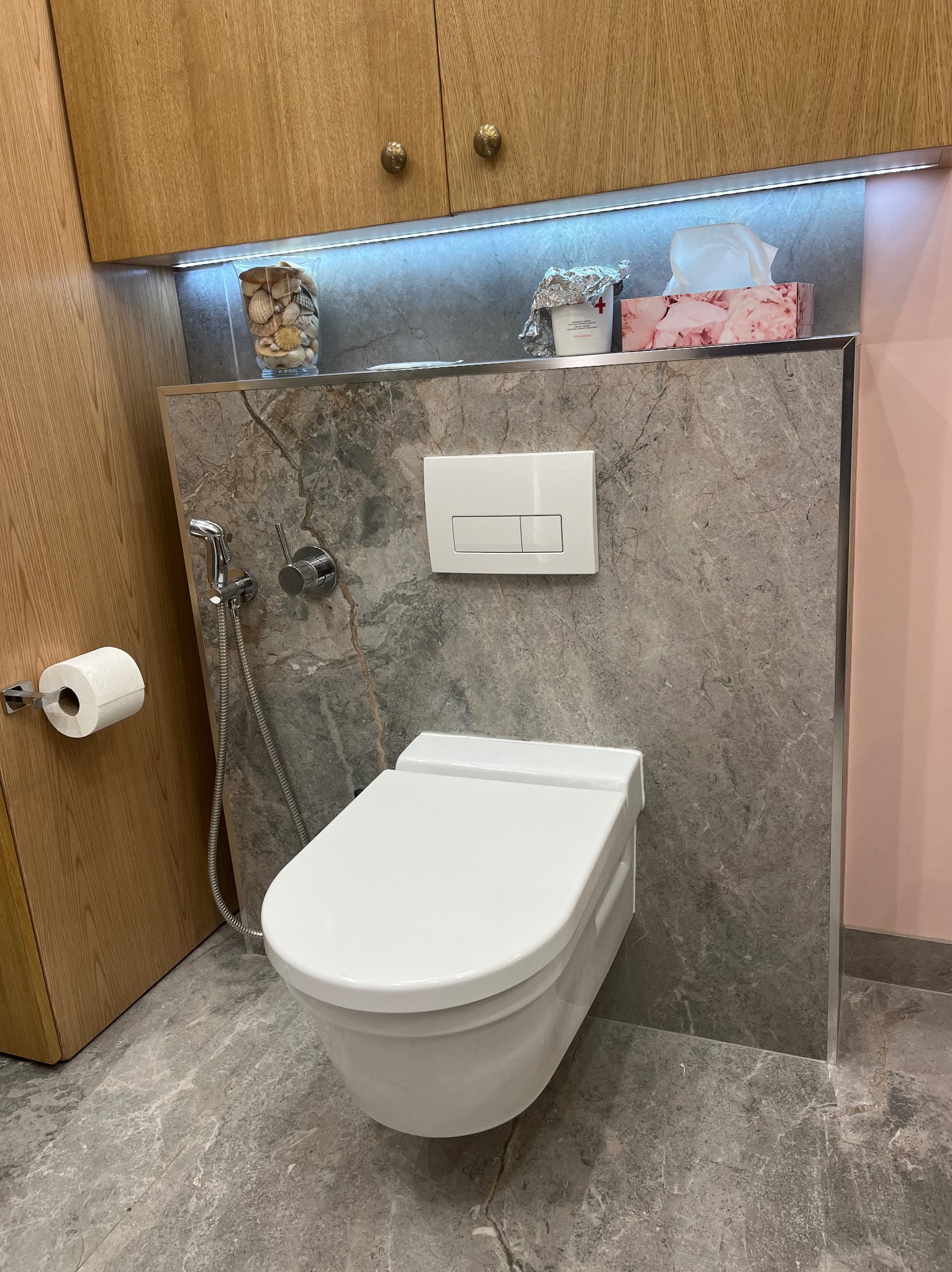 Nowoczesna toaleta podwieszana z szarą marmurową okładziną, chromowaną armaturą i drewnianymi szafkami wiszącymi, dopełniona dekoracjami z muszelek i pudełkiem chusteczek podświetlonymi ledami.