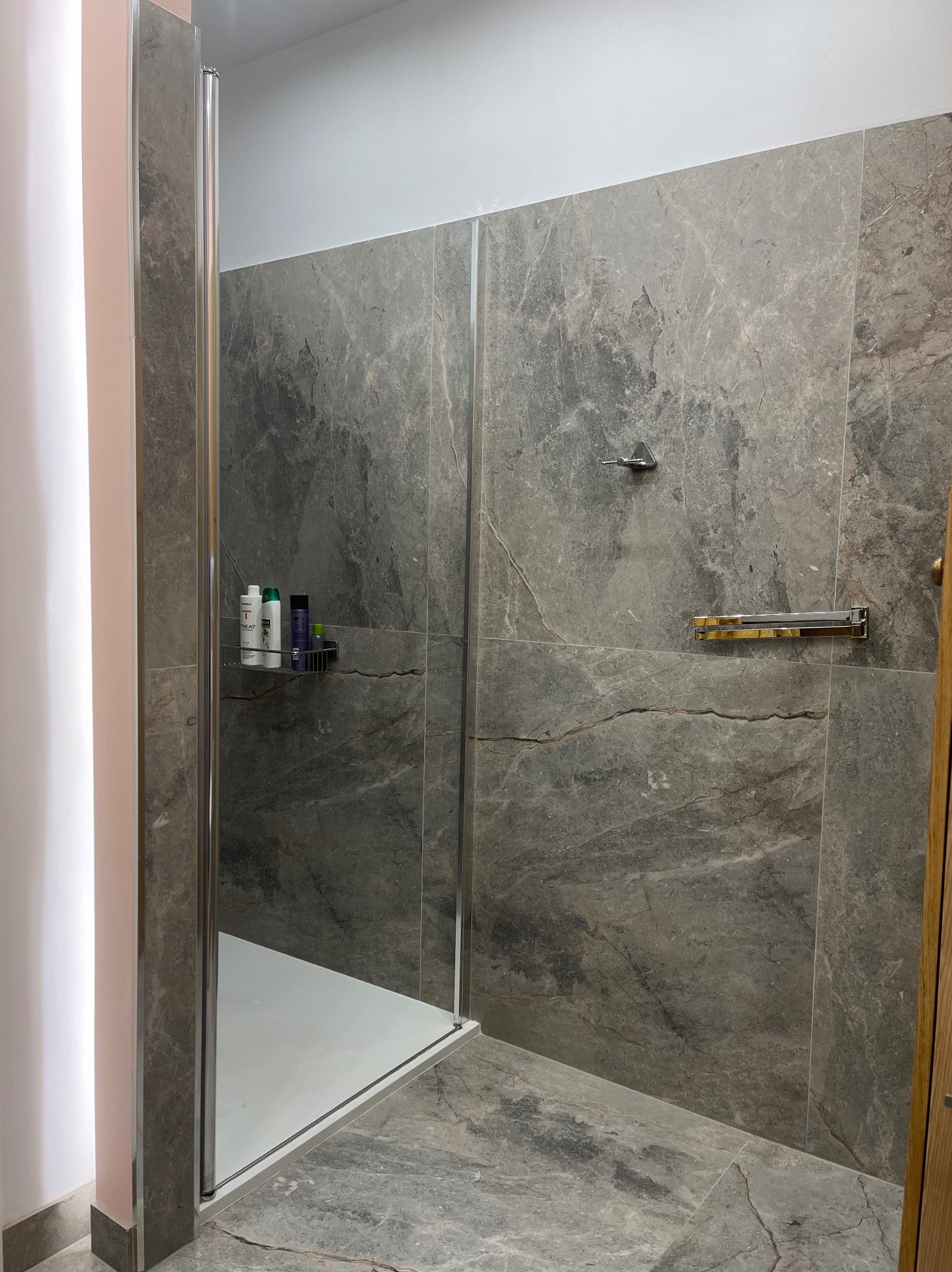 Wykończona łazienka z kabiną prysznicową, szare płytki imitujące marmur na ścianach i podłodze, metalowy wieszak na ręczniki i półka z kosmetykami pod prysznicem.