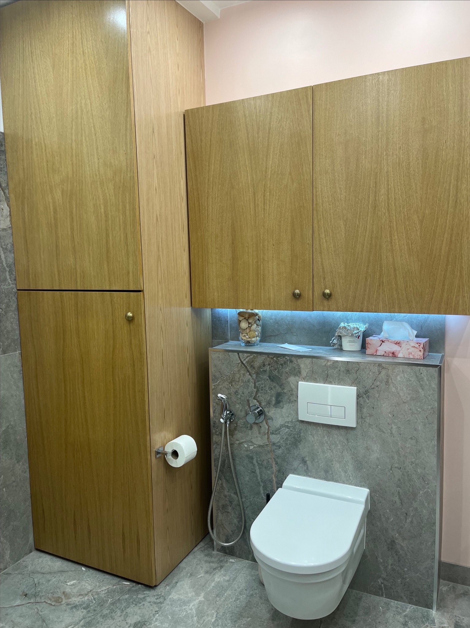 Nowoczesna łazienka z wiszącą toaletą, szarą marmurową ścianą, drewnianymi szafkami z okrągłymi uchwytami i podświetleniem LED nad półką z dekoracjami.