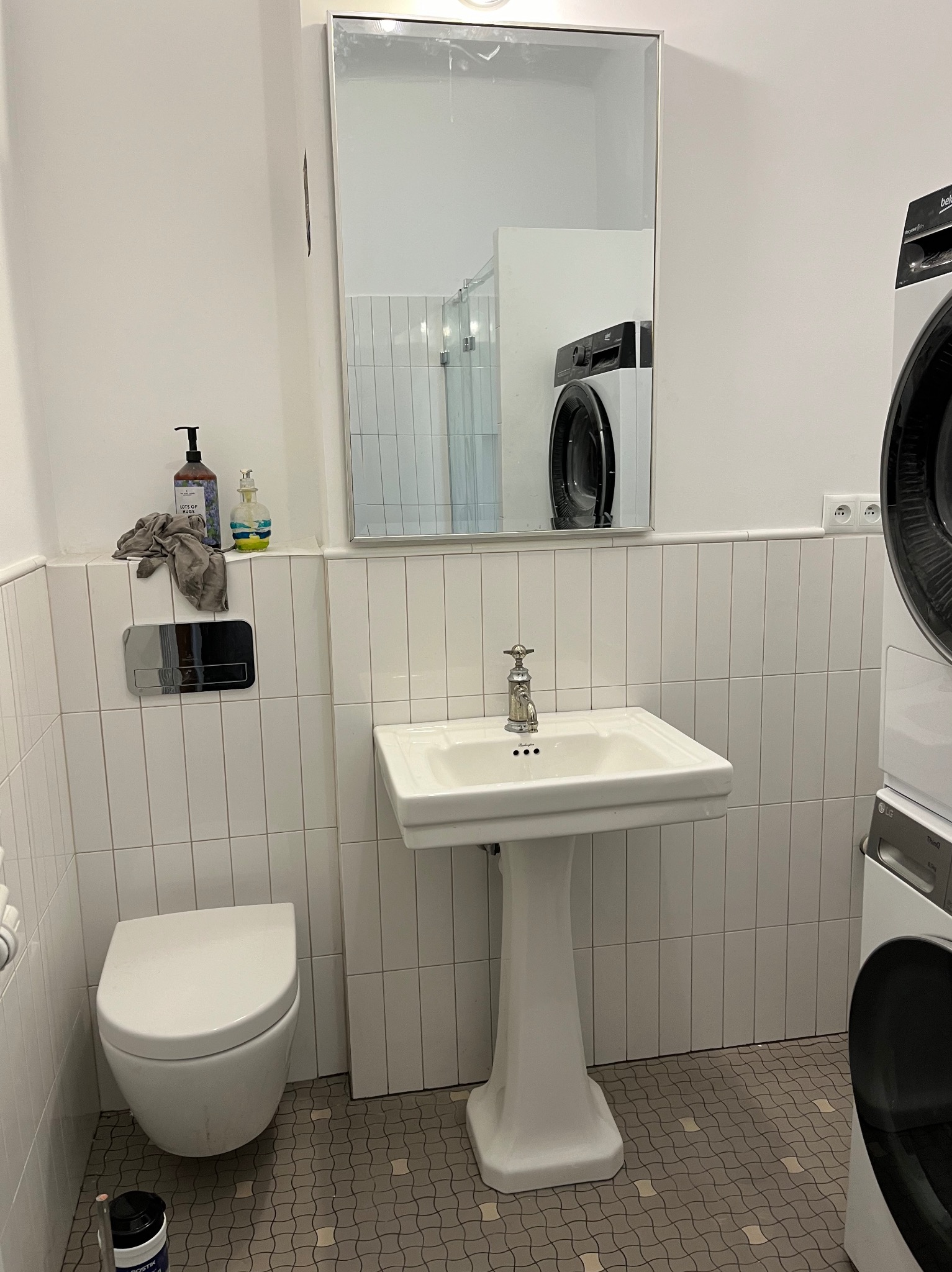 Umywalka na postumencie i wisząca toaleta w łazience wykończonej białymi, pionowymi płytkami; w lustrze odbija się pralka.