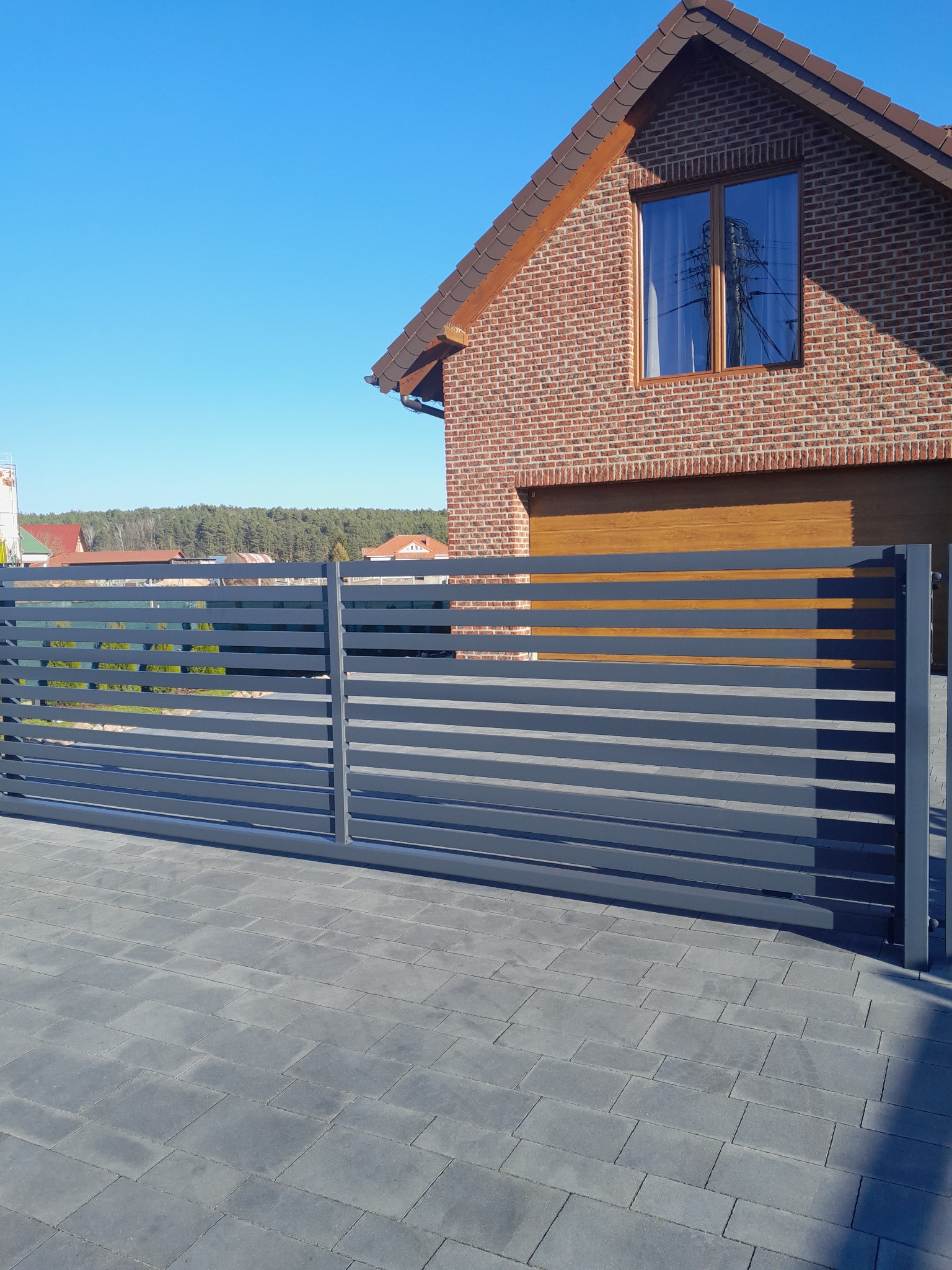 Nowoczesna, przesuwna brama wjazdowa z poziomych, szarych profili, na tle domu z cegły i kostki brukowej.