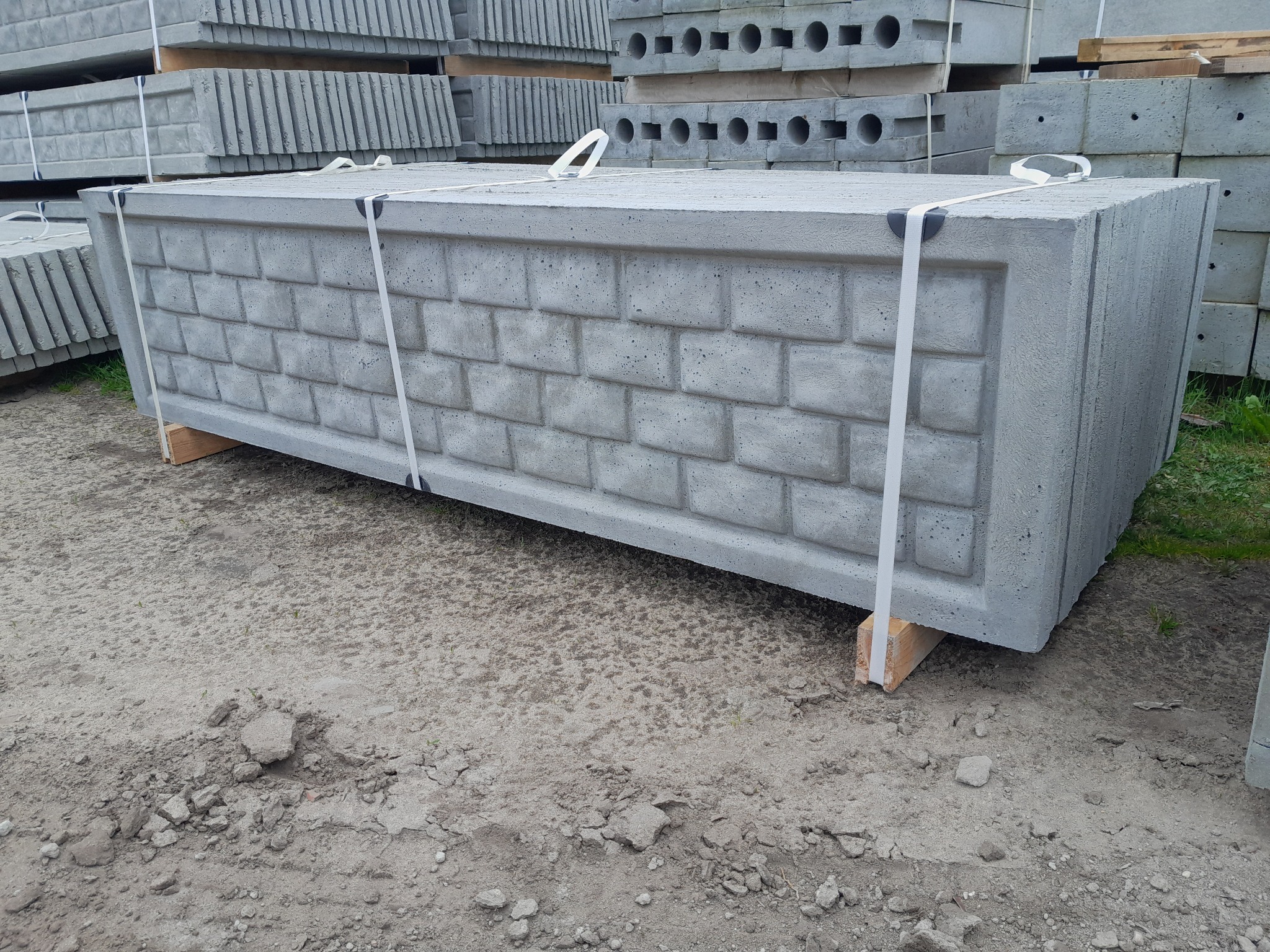 Paleta z prefabrykowanymi elementami ogrodzenia betonowego imitującego cegłę, zabezpieczona taśmami, ułożona na drewnianych podkładkach na piaszczystym podłożu.