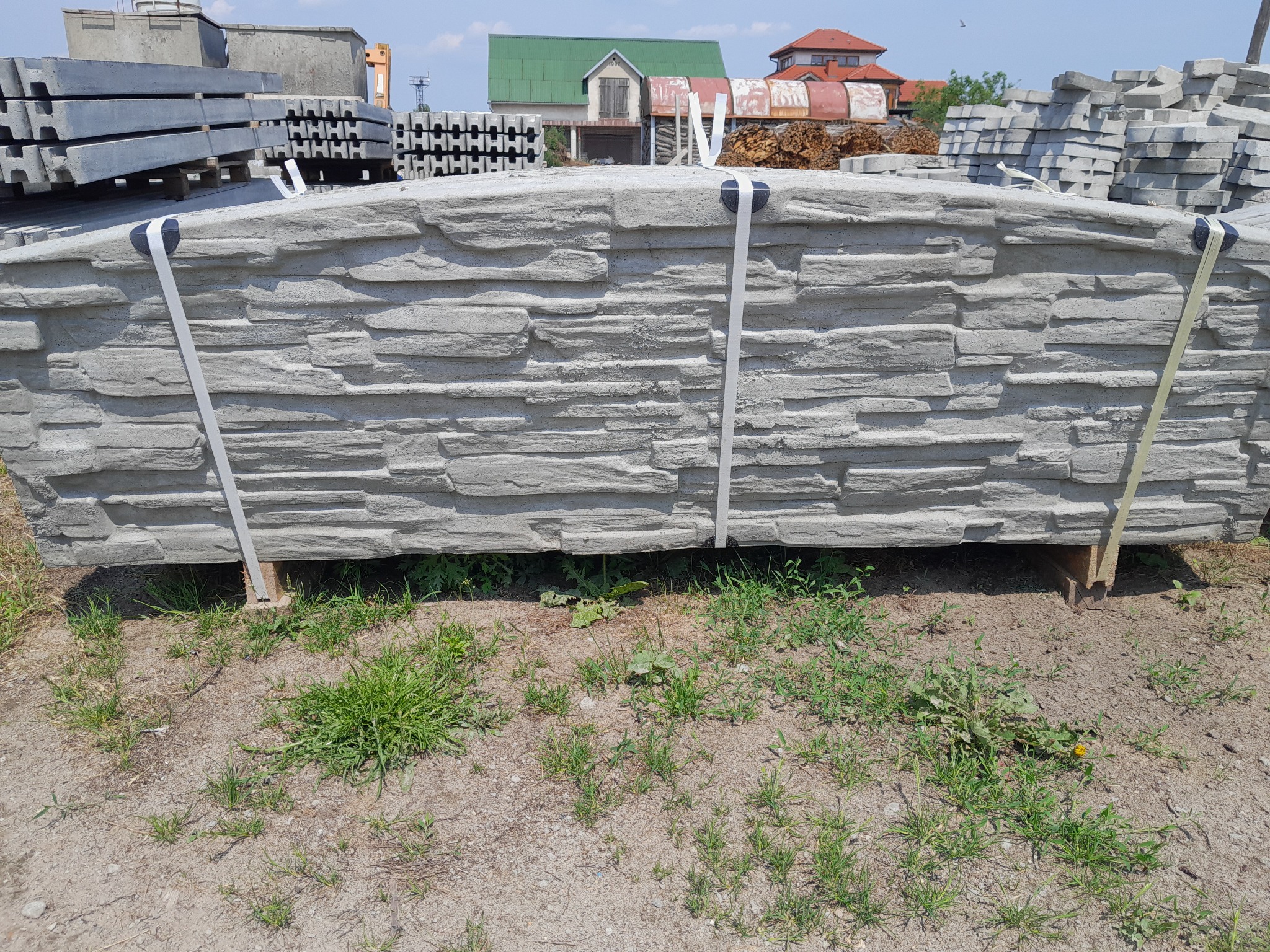 Paleta z prefabrykowanymi elementami imitującymi kamień, przeznaczonymi do budowy ogrodzenia, zabezpieczona taśmą transportową, na tle budynków mieszkalnych.