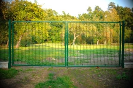 Zielona, dwuskrzydłowa brama z siatki panelowej na betonowej podmurówce, widok z zewnątrz, trawnik w tle.