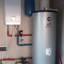 Biały piec gazowy York zawieszony na ścianie, obok pionowy, srebrny zbiornik z logo Modom Polska oraz niebieski zbiornik ciśnieniowy, połączone rurami w kolorach czerwonym i niebieskim.