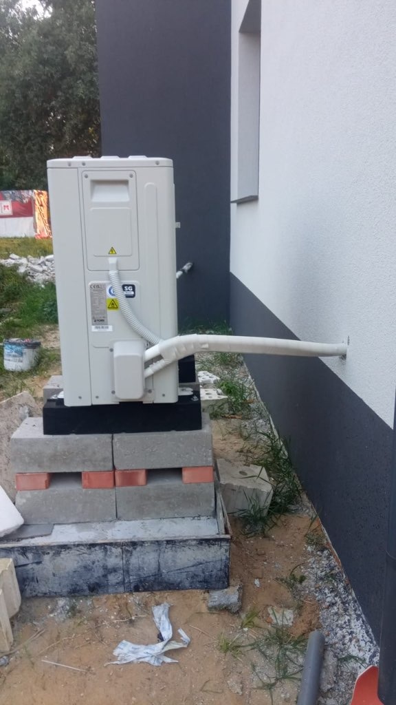 Zewnętrzna jednostka klimatyzacyjna na prowizorycznej podstawie z cegieł i bloczków, rury izolowane termicznie prowadzą do ściany budynku, widoczne naklejki ostrzegawcze i tabliczka znamionowa.